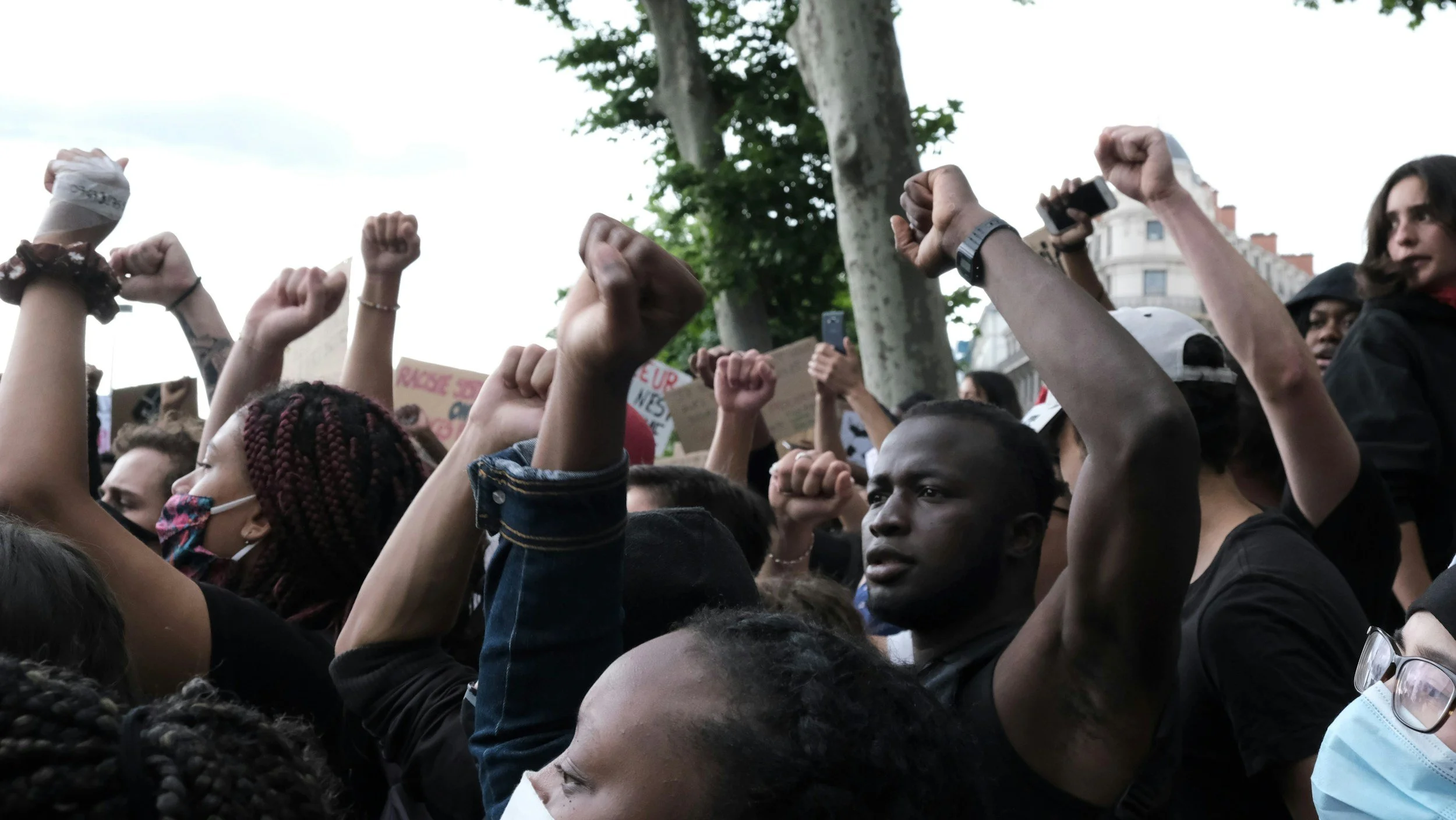 Crowd of diverse people protesting, raising fists in the air, some wearing face masks, among trees and city buildings.