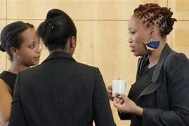 Three women in conversation, one holding a cup, in a professional setting.