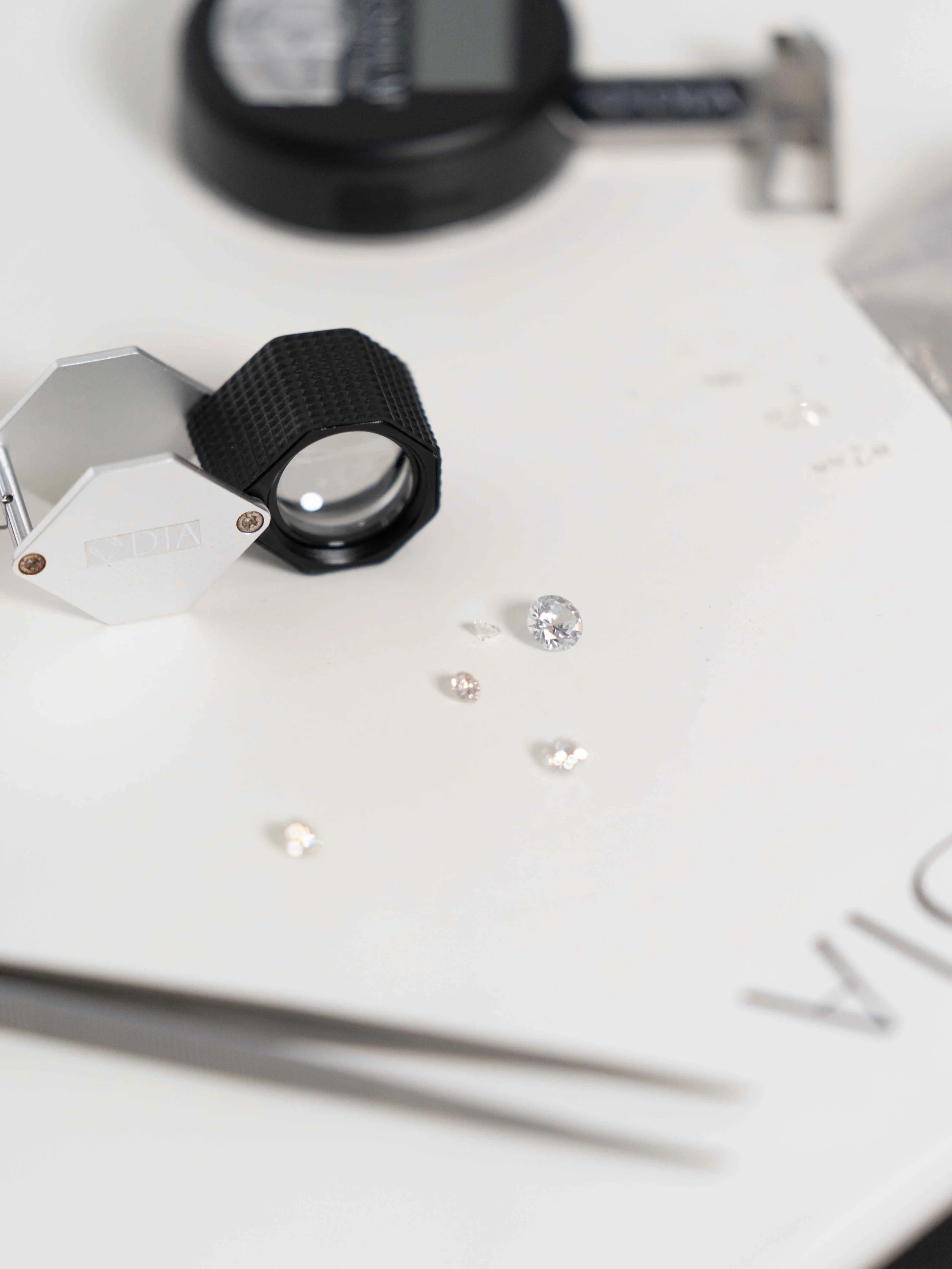 Joias sobre uma superfície branca, com uma lupa e uma ferramenta de medição ao fundo.