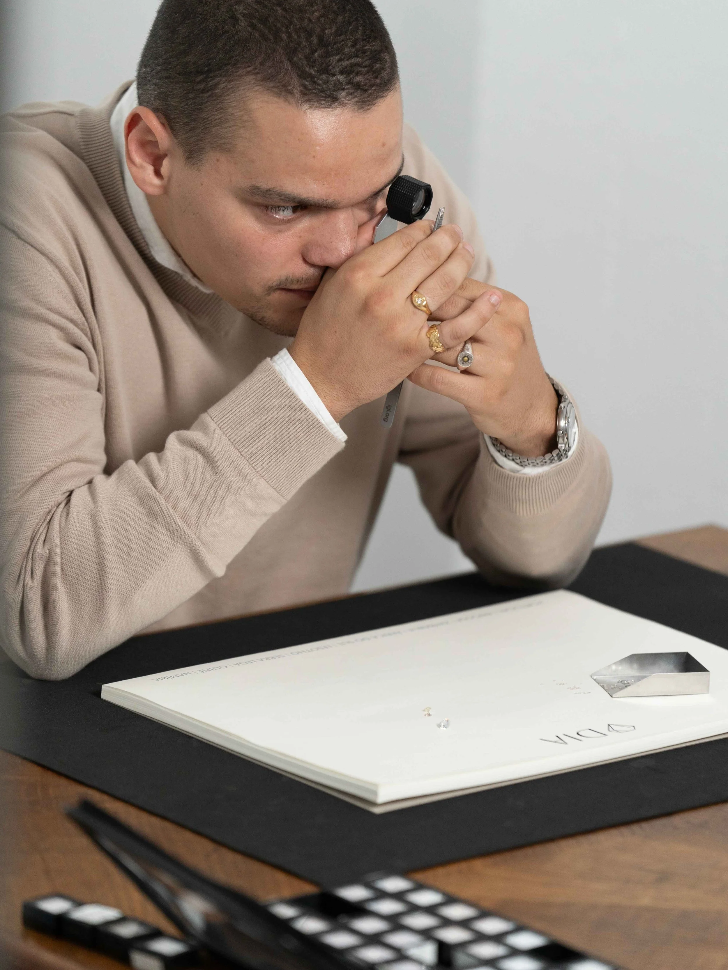 Homem analisando diamantes com uma lupa em uma mesa de madeira, papéis e ferramentas de medição ao redor.