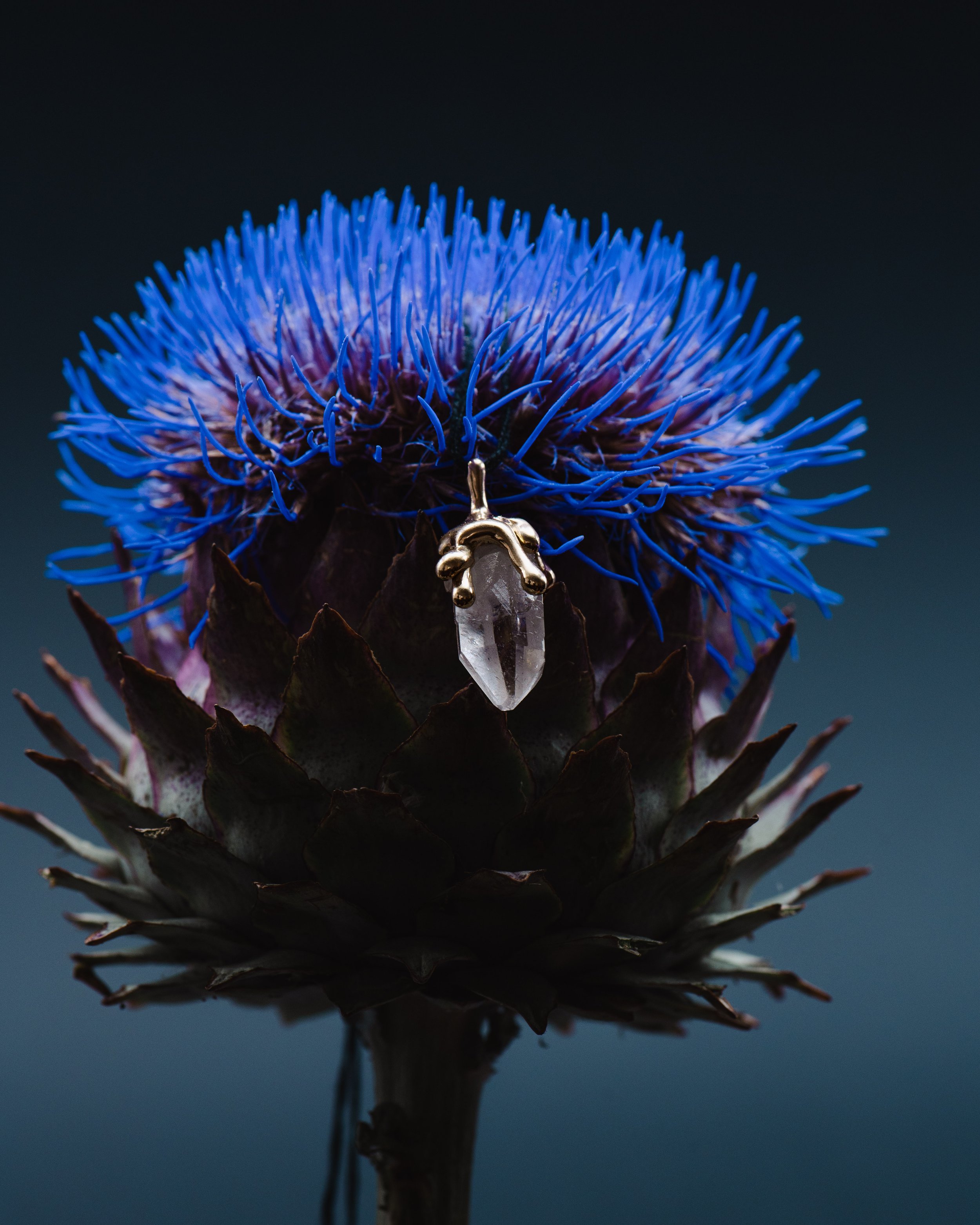 Ein blauer, stacheliger Blütenkopf einer Artischocke mit einem hängenden Kristallanhänger in Gelbgoldfassung davor