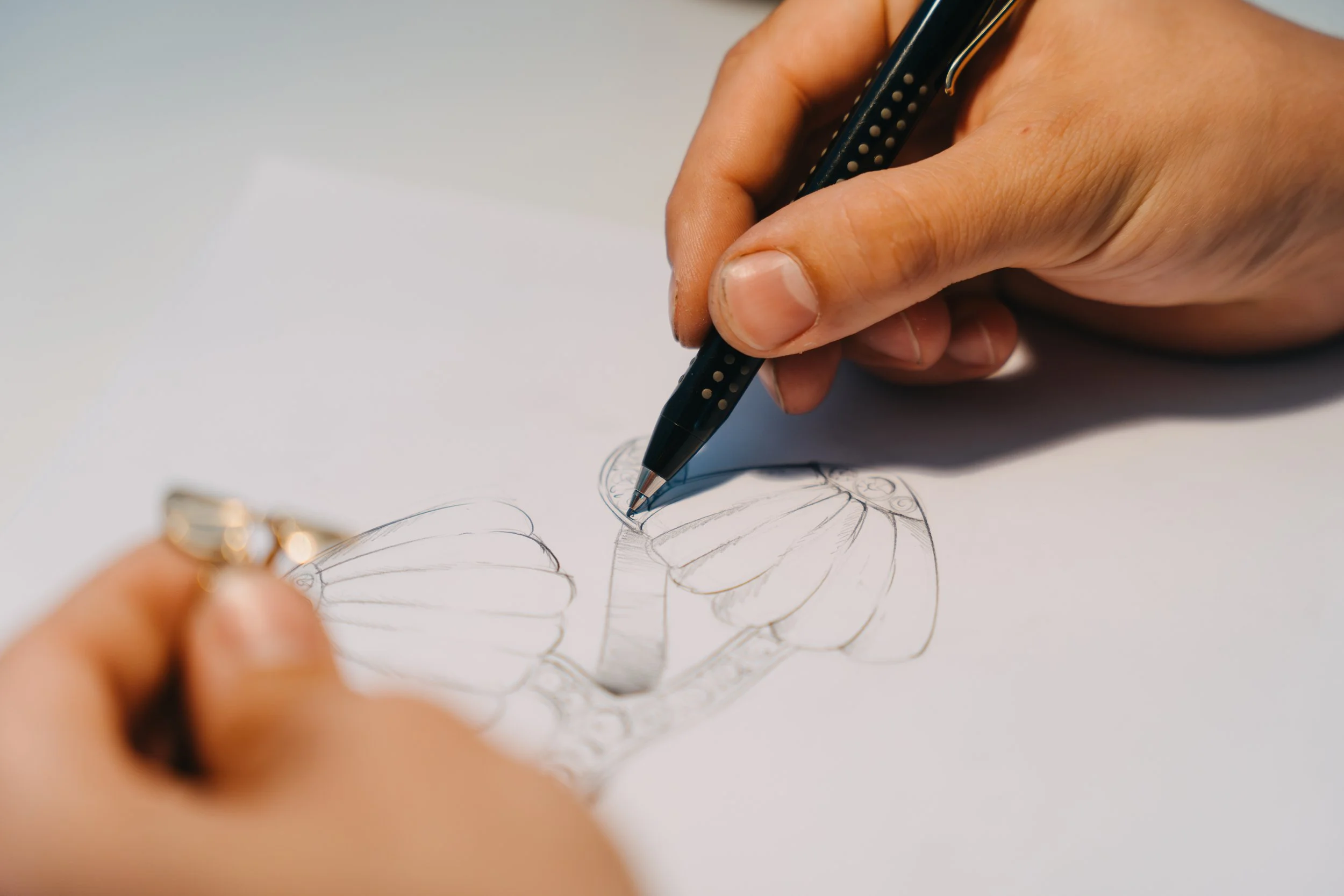 Person zeichnet eine Skizze eines Schmetterlings auf einem weißen Blatt Papier, während eine andere Hand einen Stift hält.