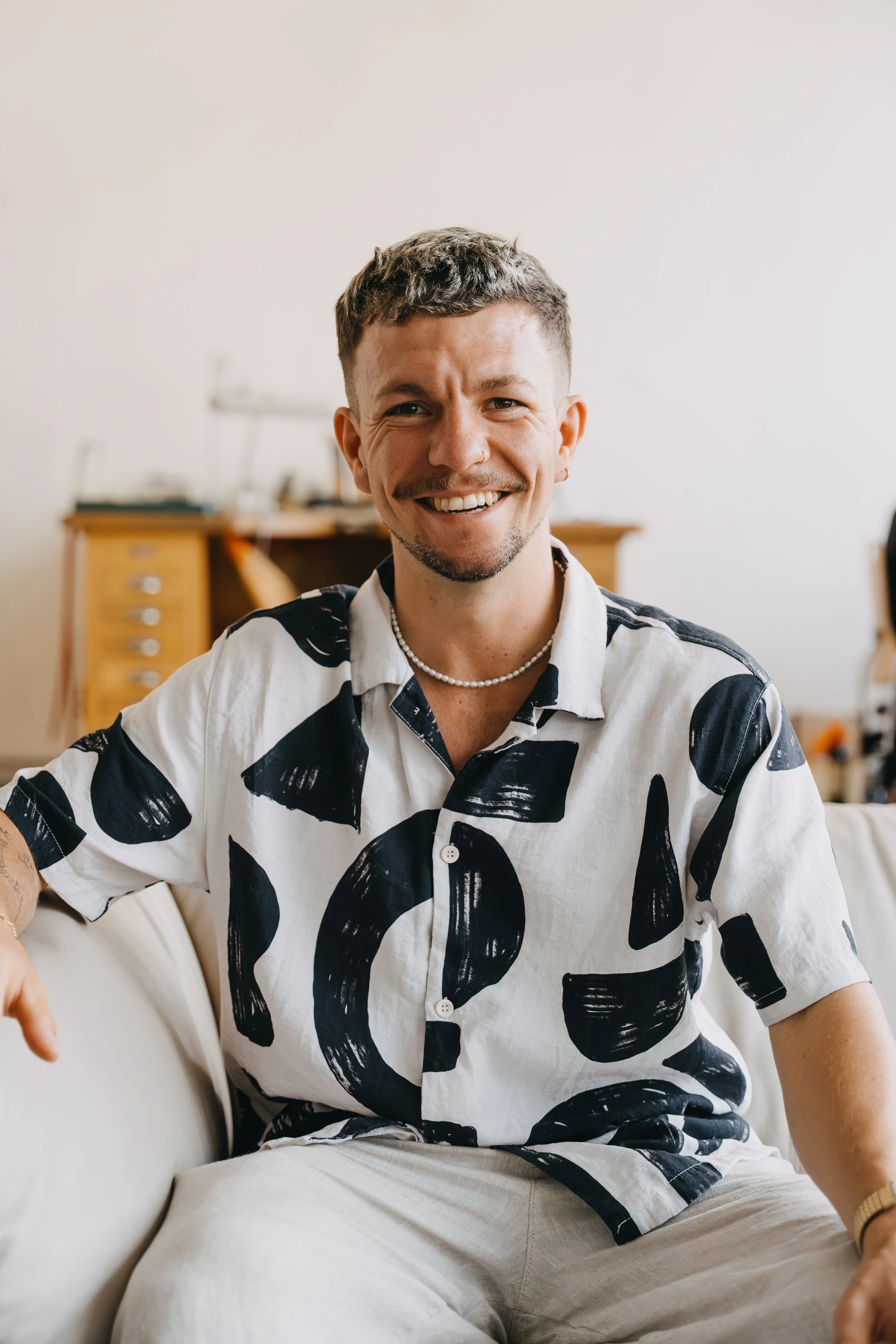 Lächelnder Mann sitzt auf einem weißen Sofa, trägt ein schwarzes und weißes Hemd mit abstrakten Mustern, eine Perlenkette, im Hintergrund ein Holztisch mit Akten.
