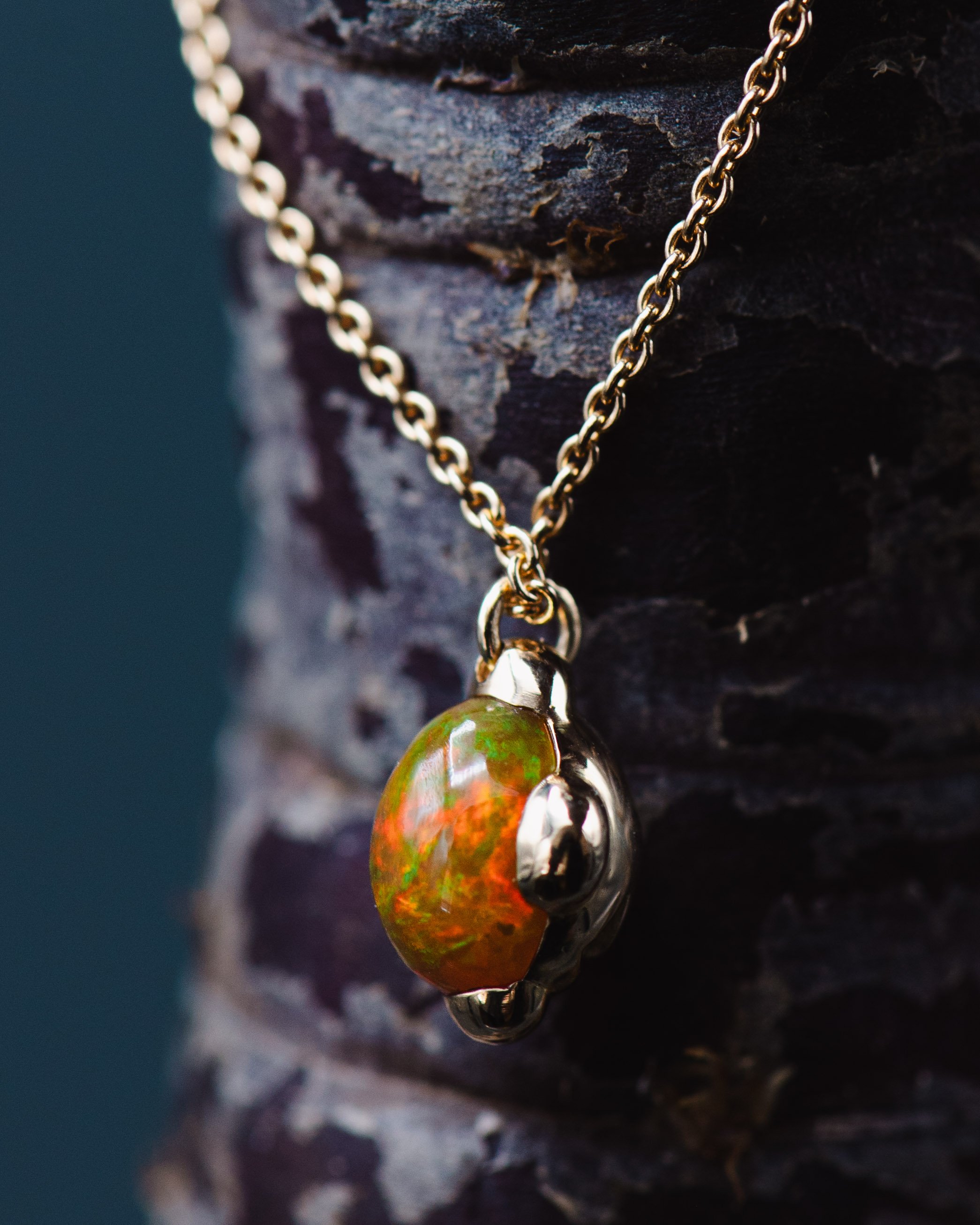 Goldene Kette mit einem Anhänger aus Opal, der um eine Metallöse befestigt ist. Der Anhänger ist oval und hat leuchtende grüne und orange Farben. Der Hintergrund besteht aus einer dunklen, rauen Baumrinde.