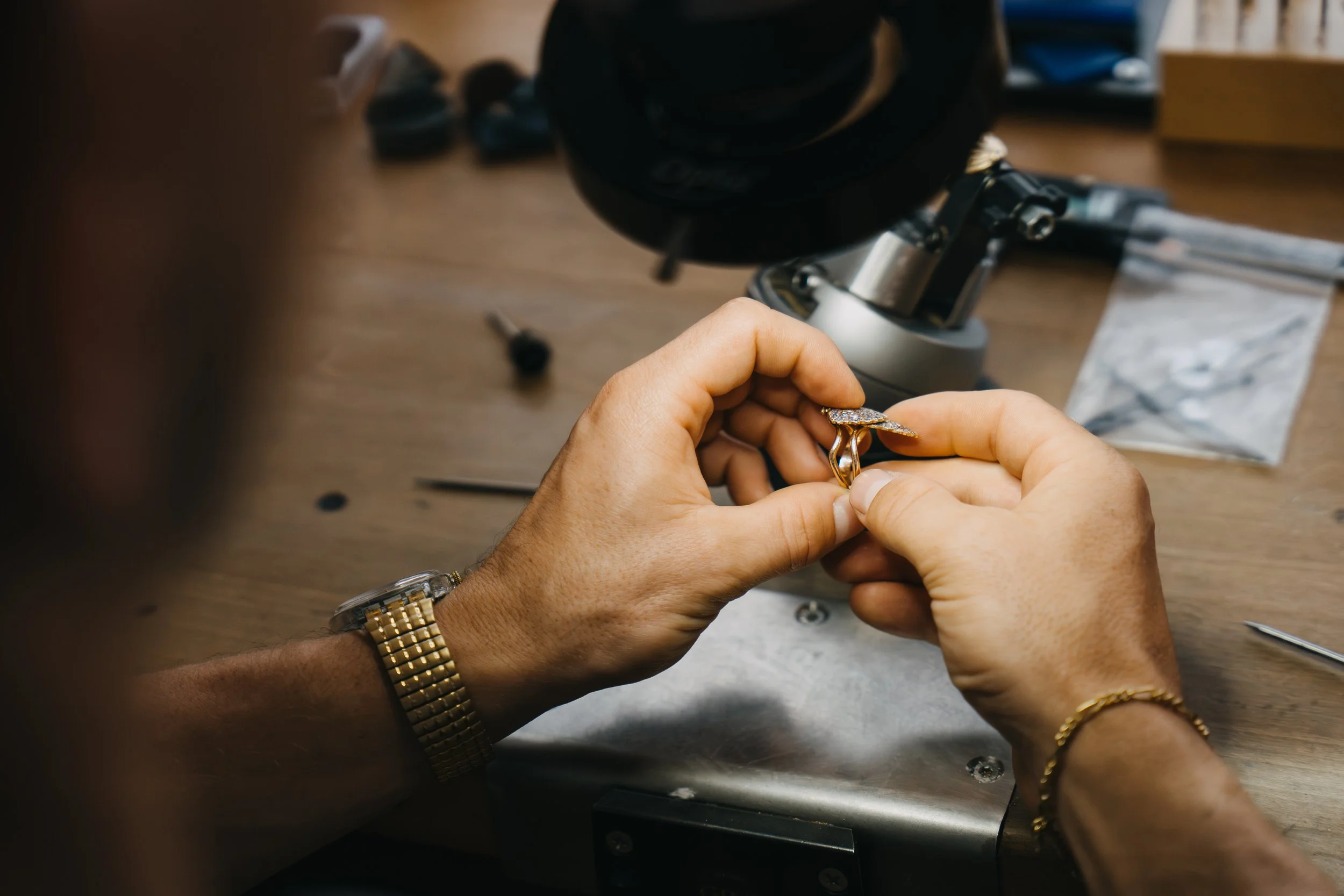 Hand arbeitet an einem goldenen Ring mit einem Mikroskop auf einem Holztisch, umgeben von Schmuckwerkzeugen und einer Papiertüte.