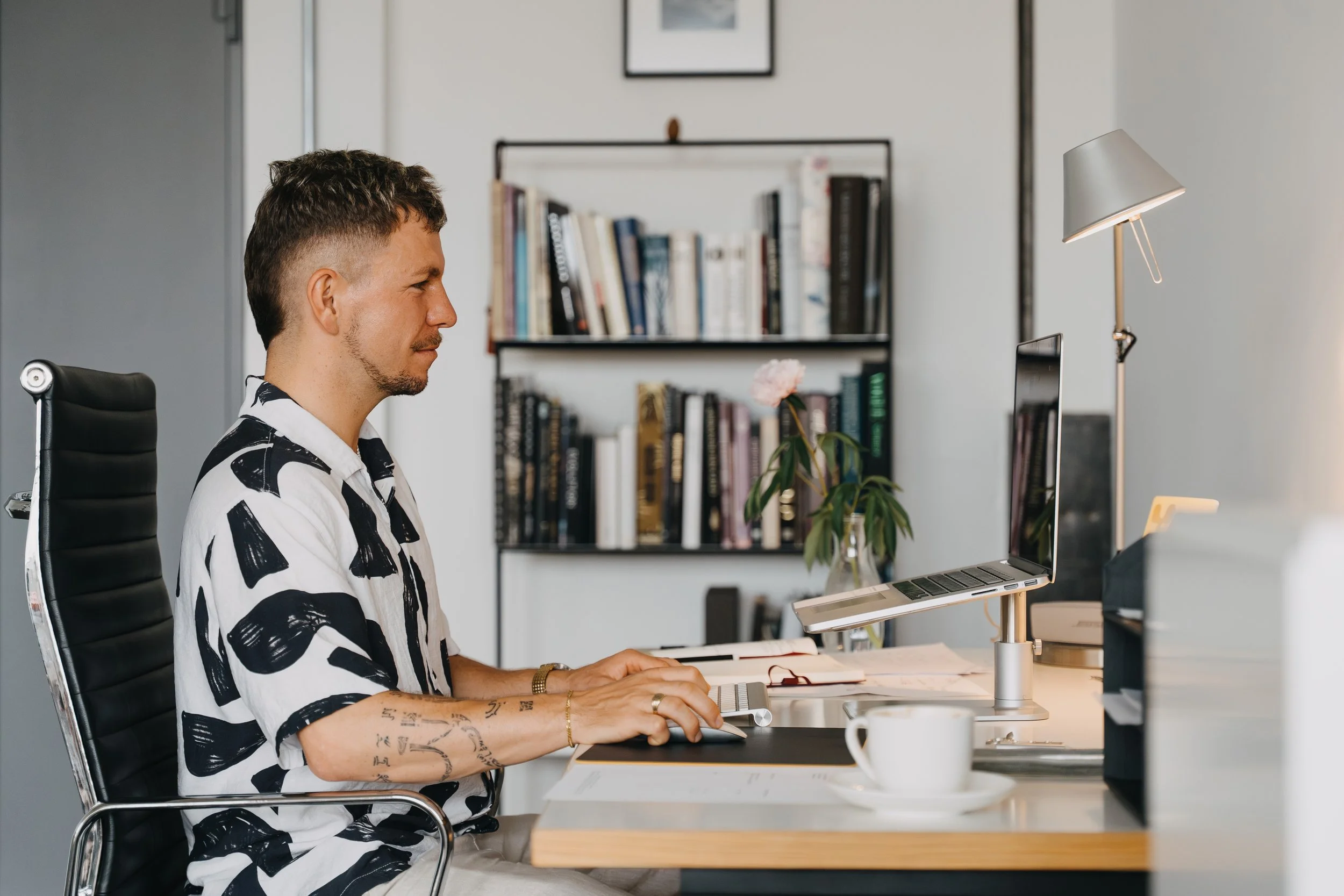 Max Sitte arbeitet an einem Schreibtisch im Büro, sitzt vor einem Laptop, mit einer Tasse Kaffee und einem Buch, im Hintergrund ein Bücherregal.