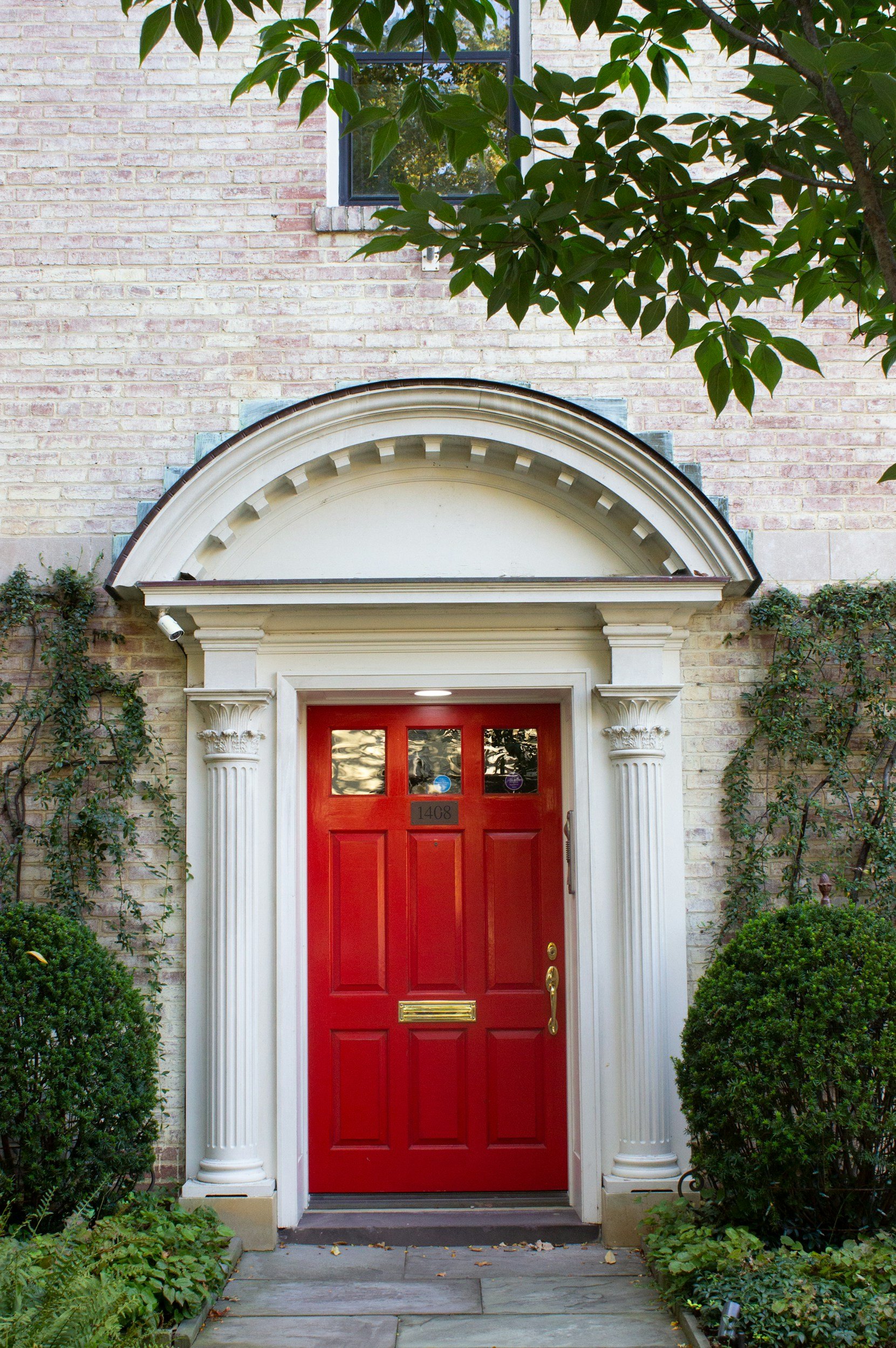 Red front door with brass hardware, white decorative columns, and an arched pediment, surrounded by green bushes and a brick building with a window above.