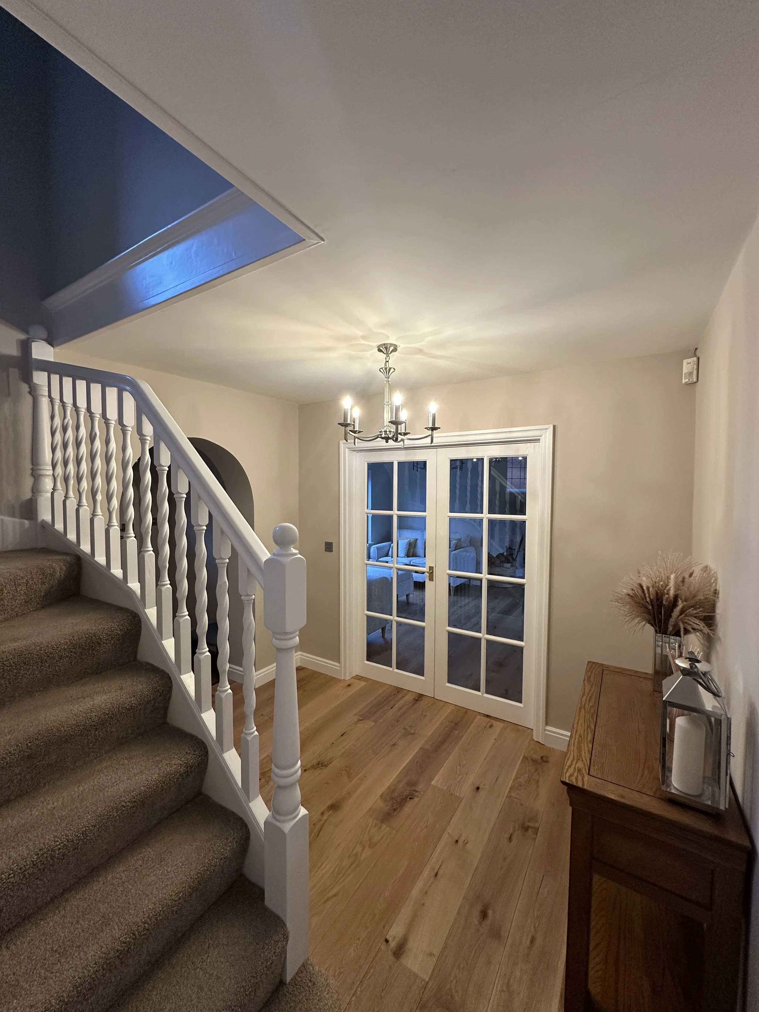 Interior view of a home's entryway with a staircase, hardwood floor, and French doors leading into another room, decorated with a chandelier and a small wooden table with dried plants.