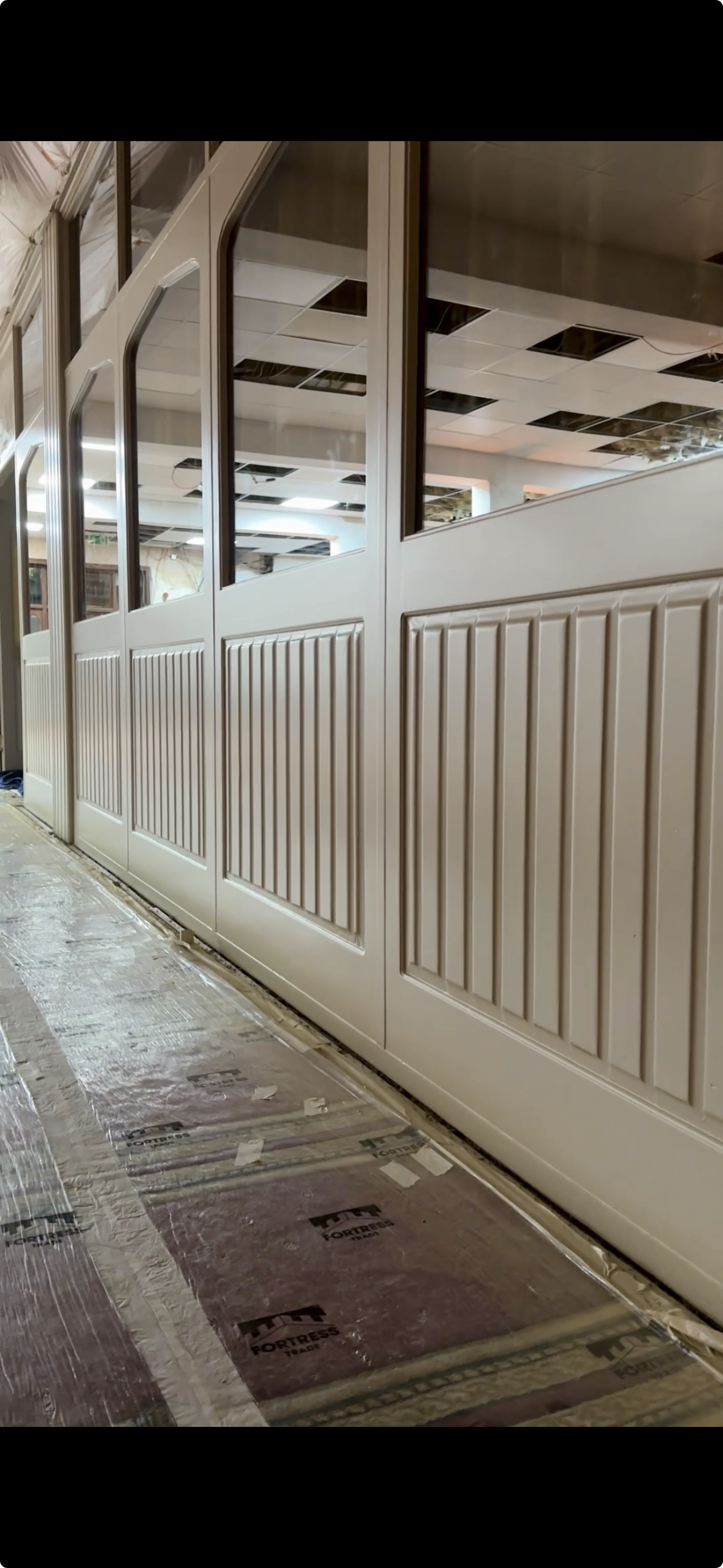 Interior view of a restaurant or cafe under construction with beige paneling and glass window dividers, floor covered with protective material.