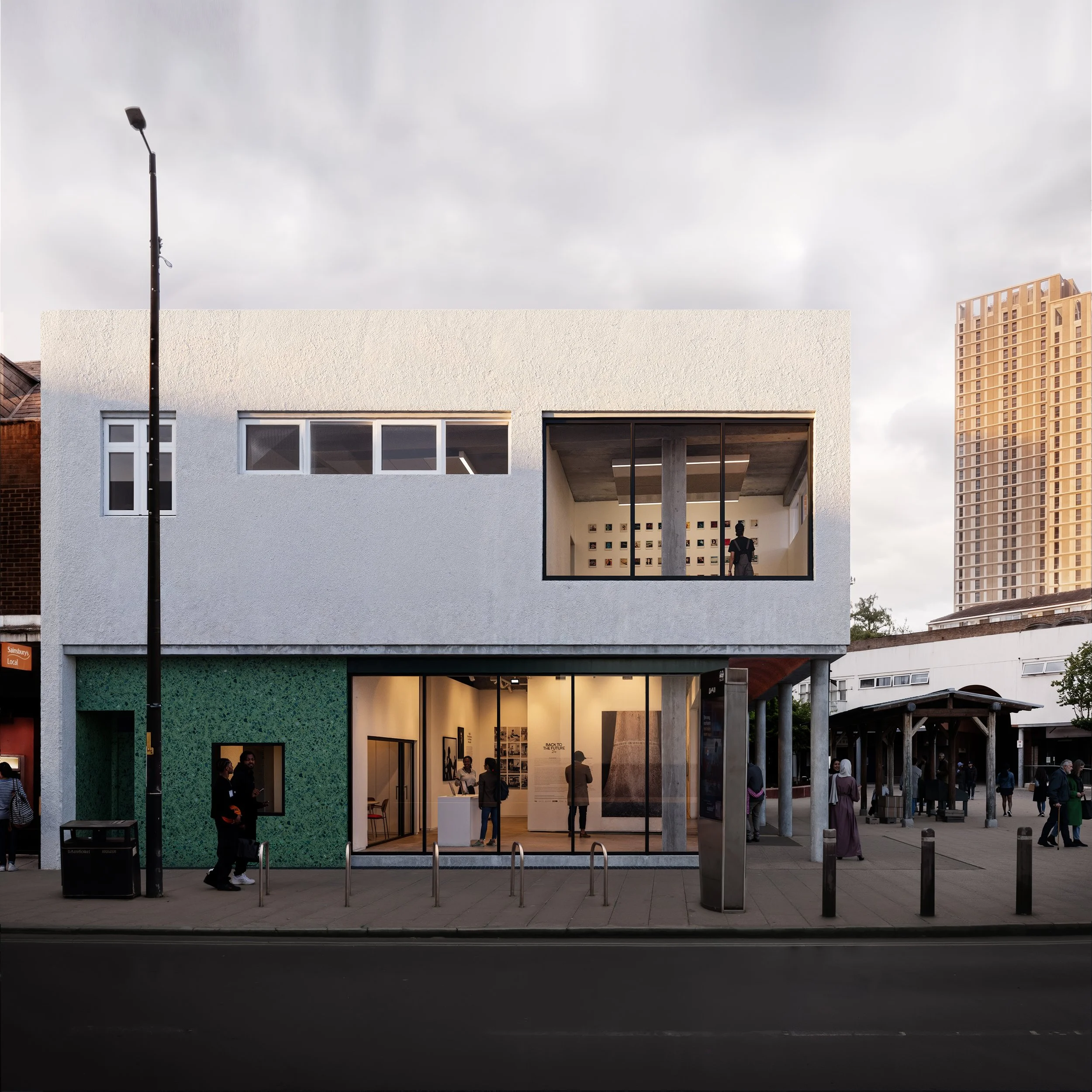 People walking in front of a modern London art venue with large windows, green wall tiling, and an open entrance. Tall buildings can be seen in the background under a cloudy sky.