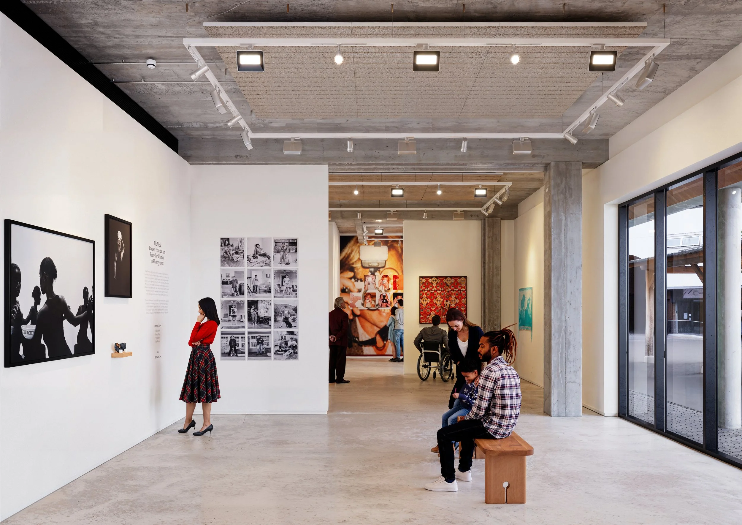 People viewing photography in a modern art gallery in Bermondsey with white walls, concrete floors and an industrial finish, some seated and some standing, with large windows showing an outdoor view.