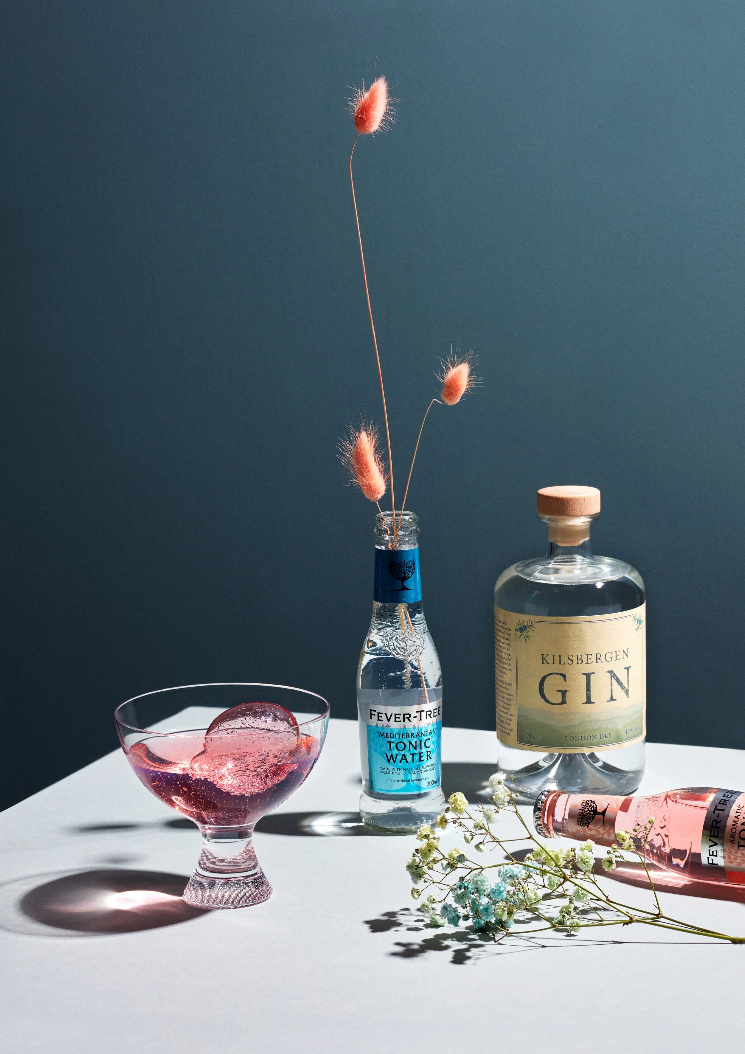 Gin och tonic flaskor står på ett vittbord med en blå bakgrund. En färdigblandad GT står på bordet. Ur en tonicflaska sticker det upp små rosa växter, harsvansar.