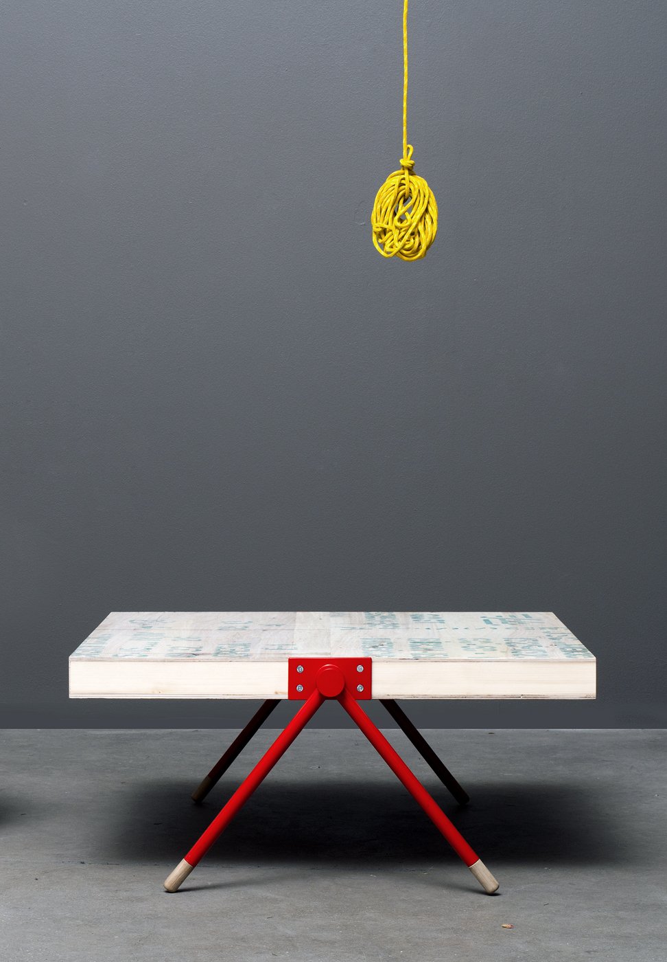 Minimalist table with red metal legs and wooden top, yellow rope hanging above, gray wall background.
