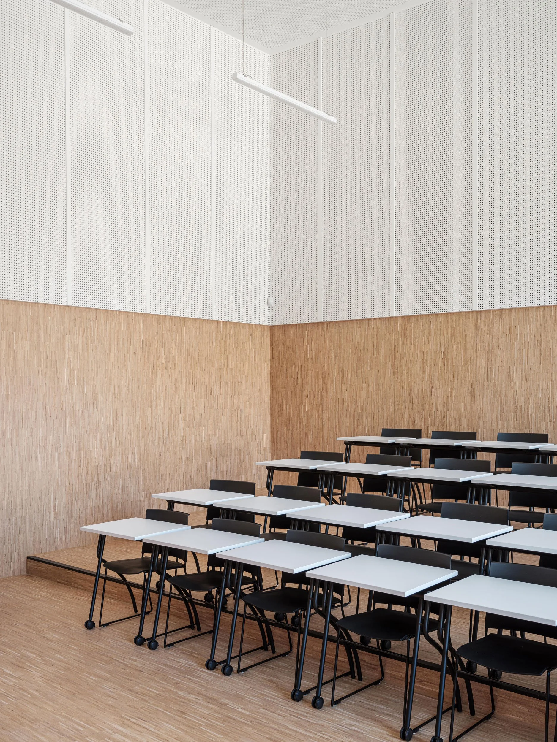 Empty classroom with rows of black chairs and white desks, high ceilings, light-colored wooden walls, and suspended fluorescent lighting.