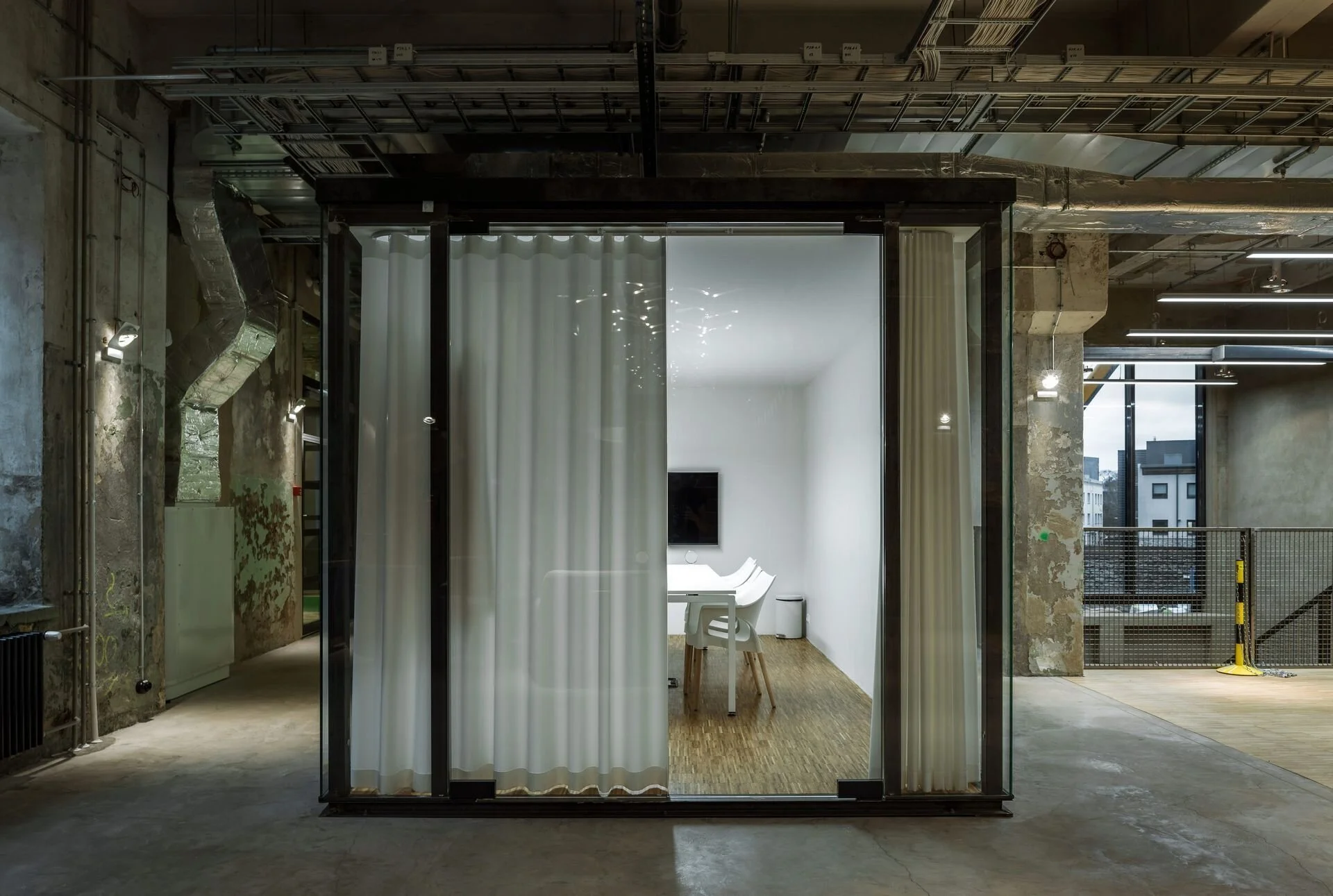 Modern minimalist office space with glass walls, white curtains, a table, and chairs, set in an industrial-style building with exposed ducts and concrete walls.