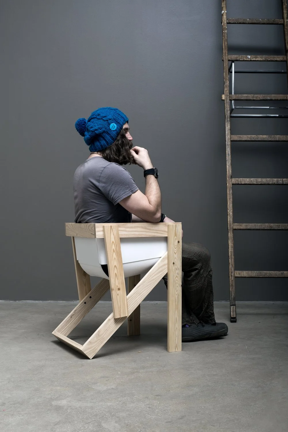Man with blue beanie sitting in unique wooden chair next to ladder on concrete floor.