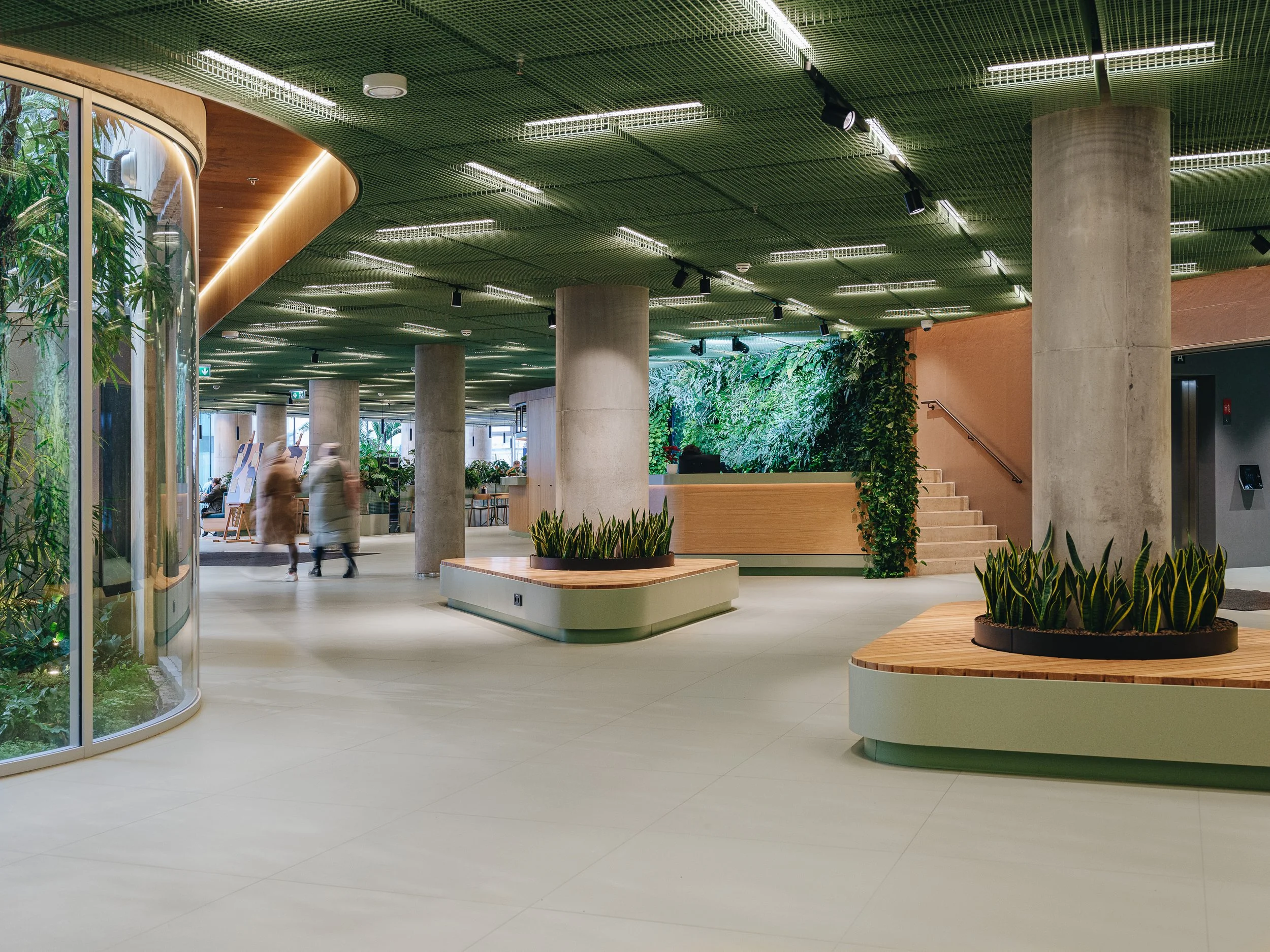 Modern interior space with green plants, large columns, and people walking. Features staircase and elevator on the right.