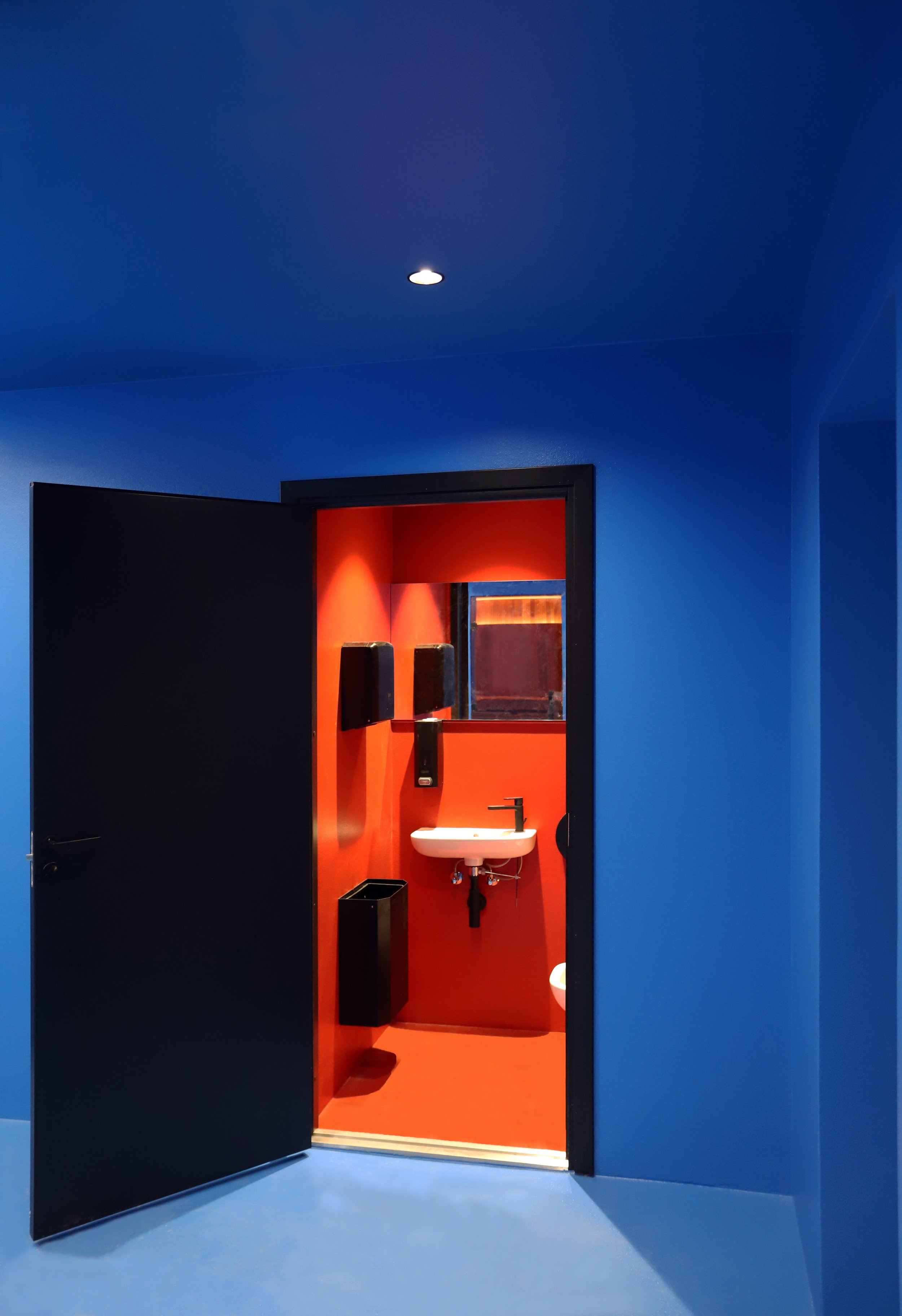 Open door to a brightly lit red bathroom with a sink and mirror, set within a contrasting blue room.