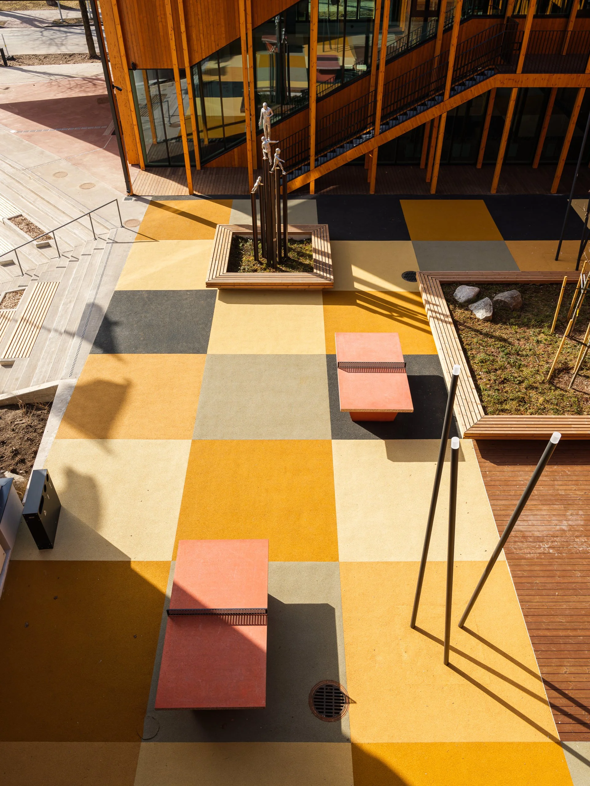 Modern outdoor plaza with colorful patterned concrete tiles, benches, planters, and an abstract sculpture near a building with large windows and wooden accents.