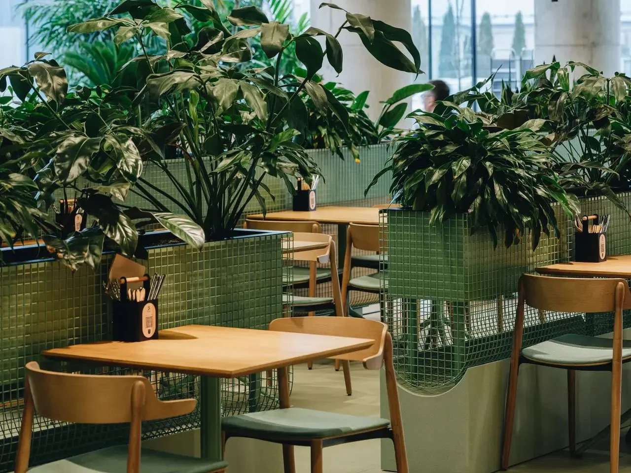 Indoor café seating area with wooden tables and chairs, surrounded by lush green plants in modern planters.