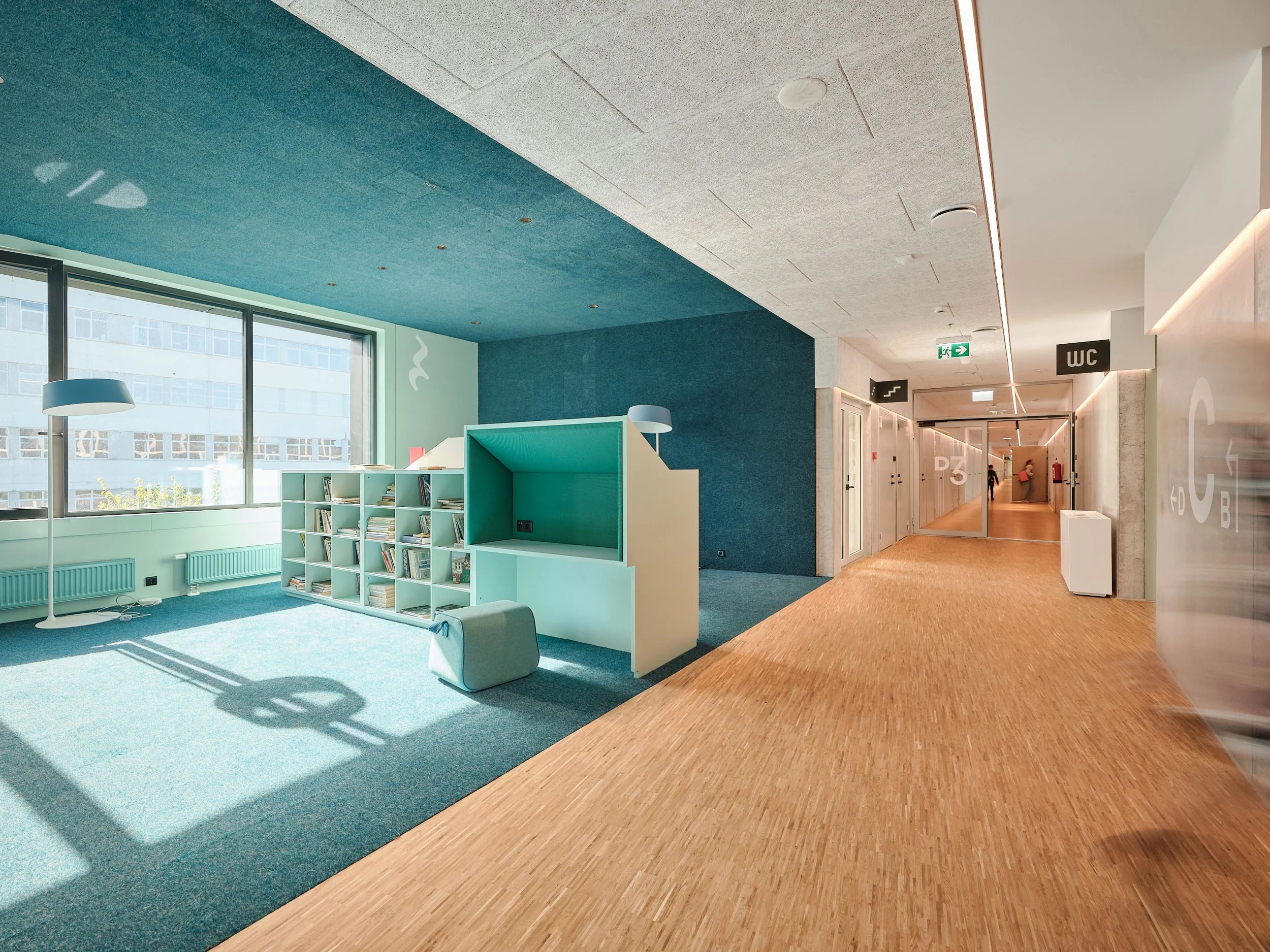 Modern office interior with a bright teal and wooden color scheme, featuring a reading area with bookshelves and chairs, large windows, and a corridor leading to rooms with labeled doors.
