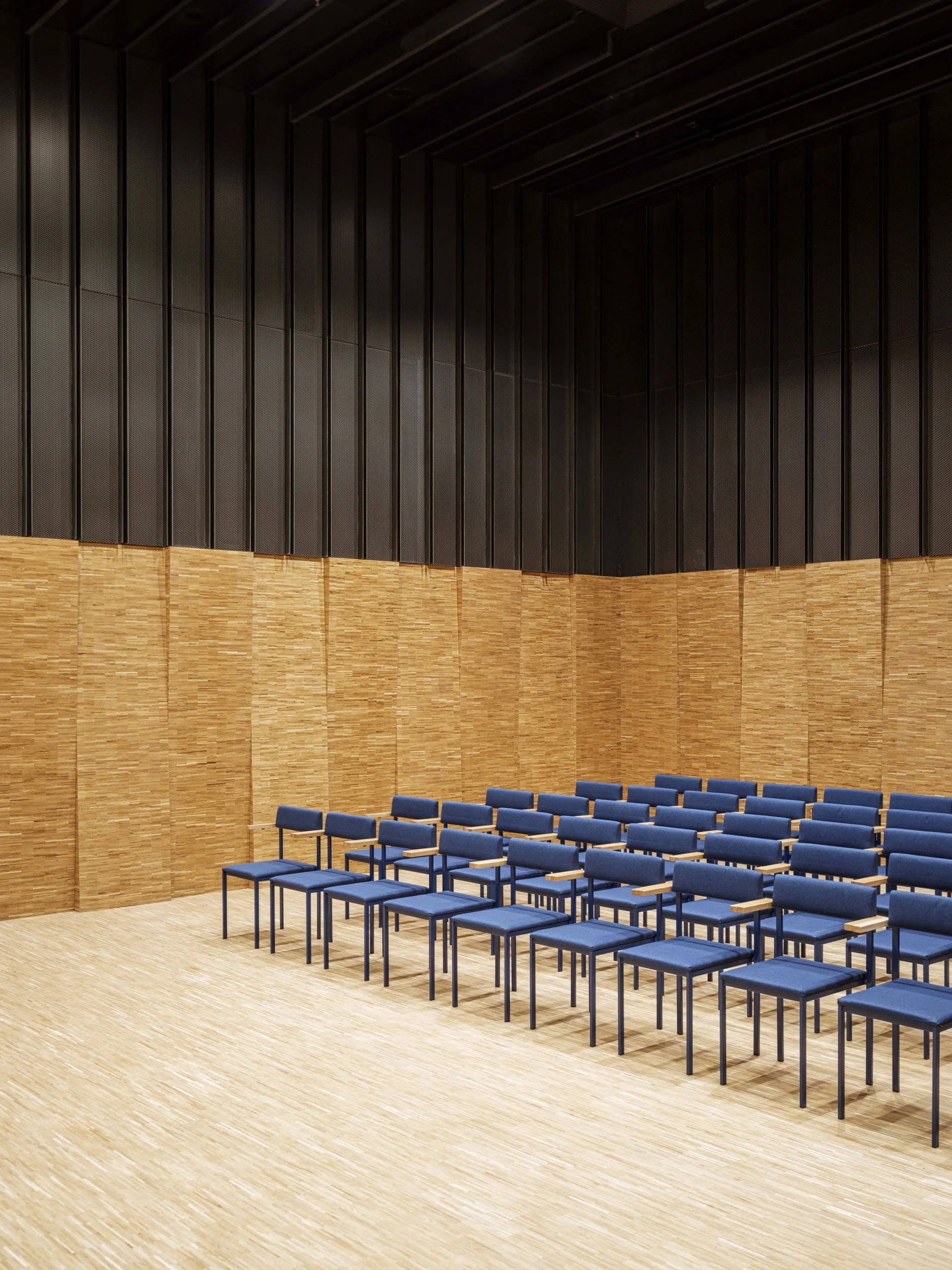 Modern lecture hall with wooden panels and rows of blue chairs.