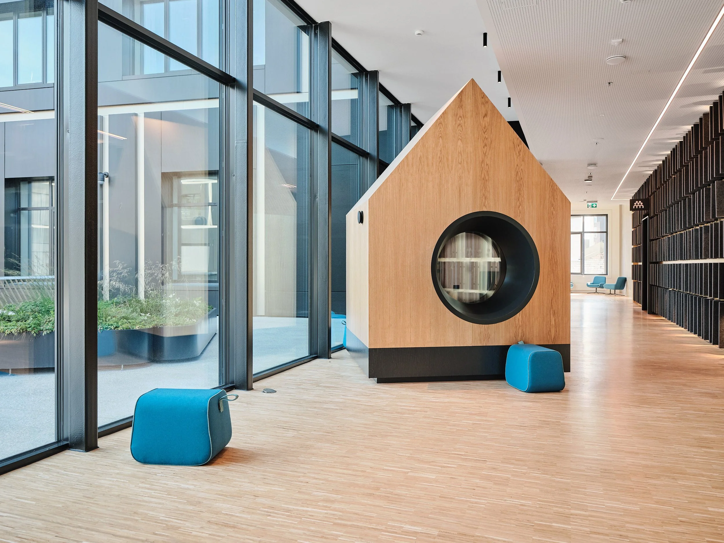 Modern office lobby with large windows, wooden flooring, a unique wooden structure with a circular opening, and blue seating cubes.