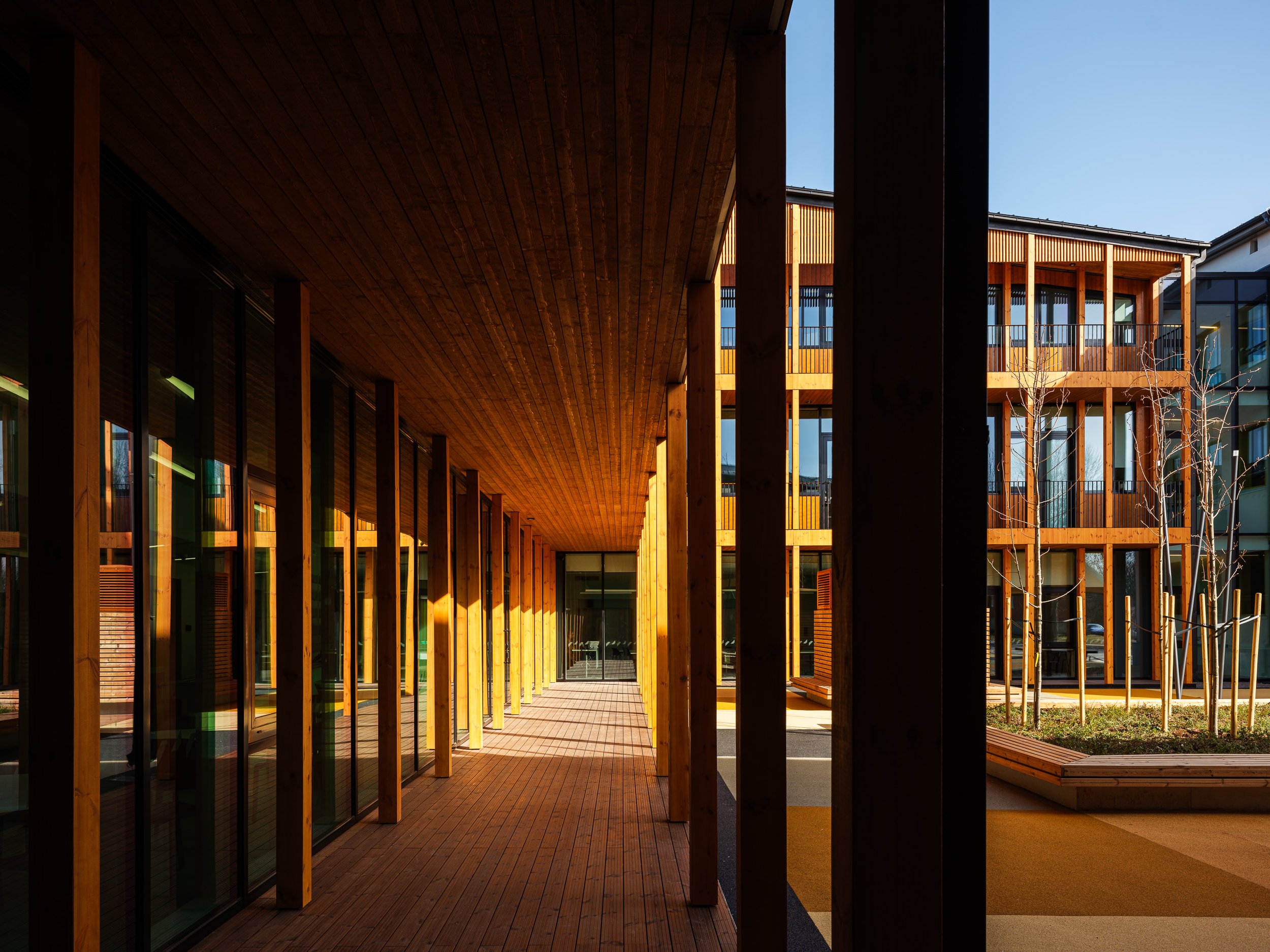 Modern wooden architectural building with glass windows, featuring a covered walkway with vertical wooden beams and an open courtyard illuminated by sunlight.