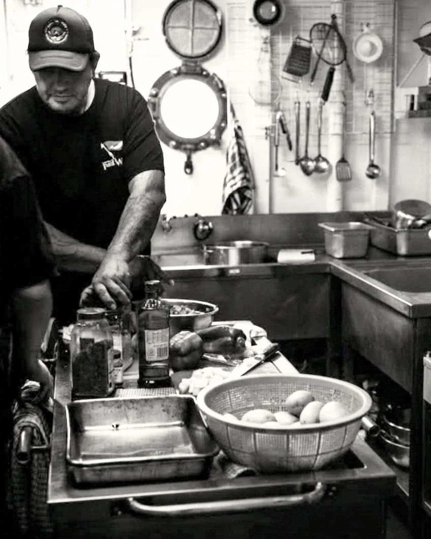 A man preparing food in a kitchen with cooking utensils hanging on the wall behind him, with a bowl of eggs, canned goods, and various vegetables on the counter.