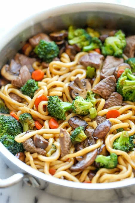 Cooked stir-fry with beef, broccoli, carrots, mushrooms, and noodles in a metal pan