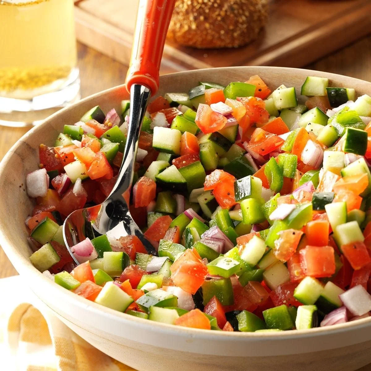 A bowl of freshly chopped vegetable salad with cucumbers, tomatoes, and onions, with a fork inside the bowl, on a wooden surface, alongside a glass of beverage and a loaf of bread in the background.