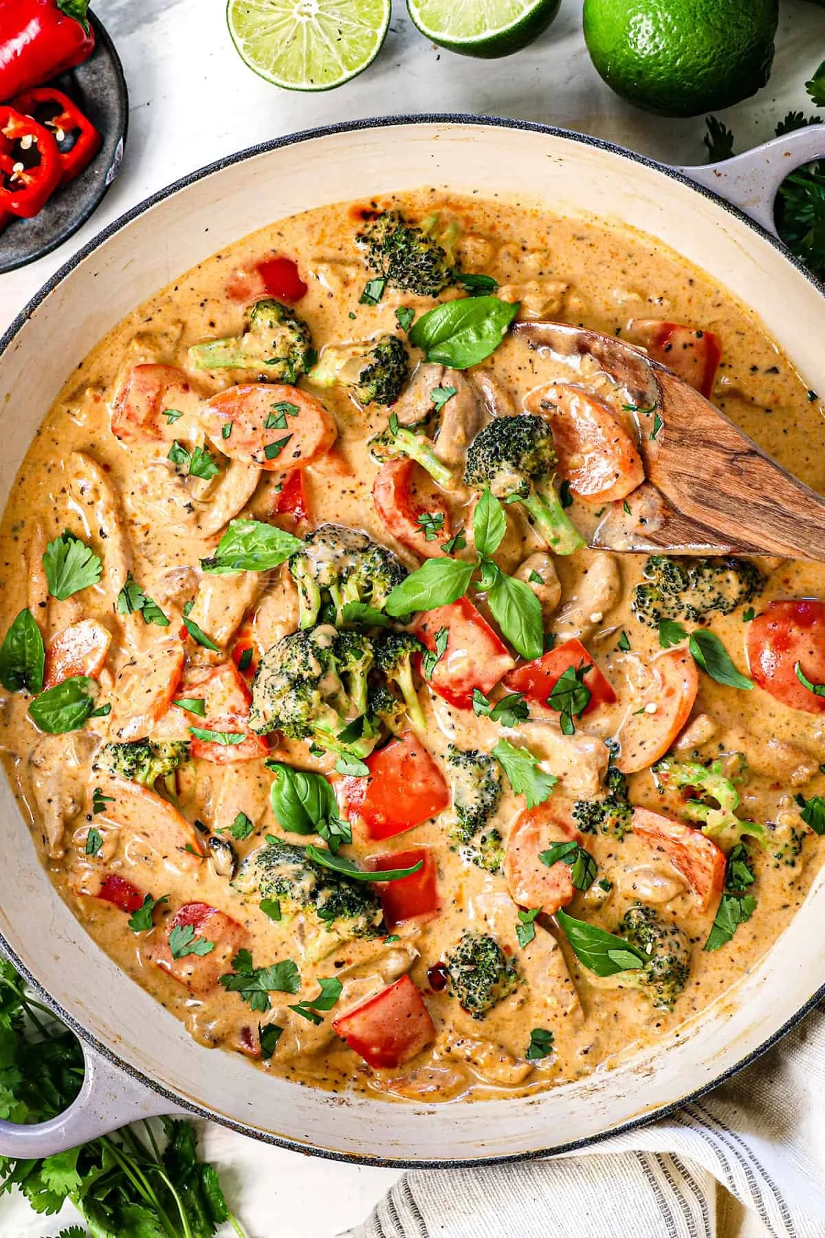 Creamy vegetable curry with broccoli, tomatoes, and cilantro in a white bowl. Sliced limes and whole lime on a white surface surround the bowl.
