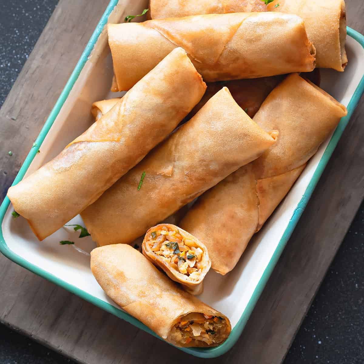 A bowl of golden fried spring rolls, with one cut in half showing a vegetable filling, on a wooden surface.