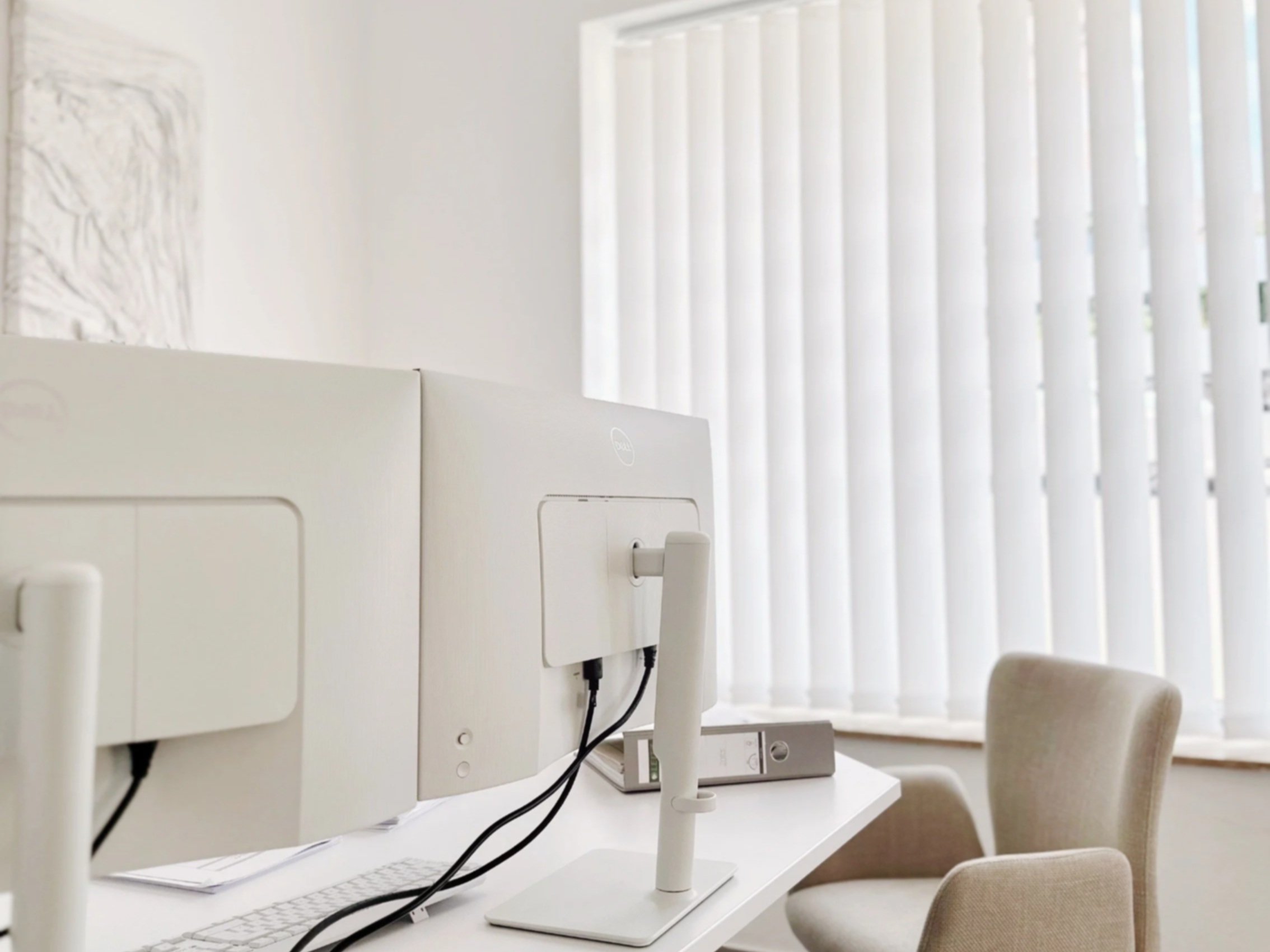 Ein heller Büroarbeitsplatz mit zwei weißen Computermonitoren, einem Computer, einer Tastatur, einem weißen Ständer, einem beige-grauen Bürostuhl und einem Fenster mit vertikalen weißen Jalousien.