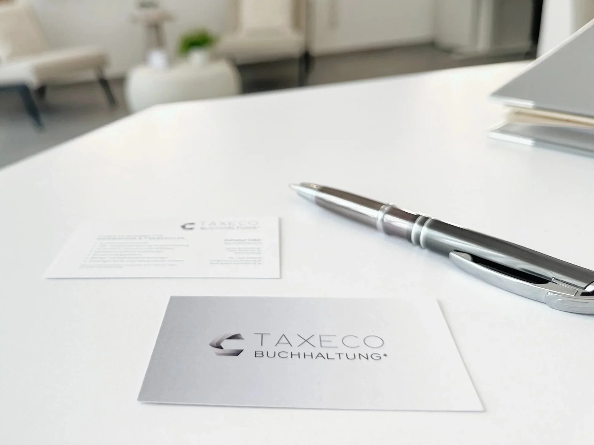 Ein Whiteboard mit einem Stift, ein Blatt Papier mit 'TAXECO BUCHHALTUNG' Logo, und weiteres Papier auf einem weißen Tisch in einem modernen Büro.