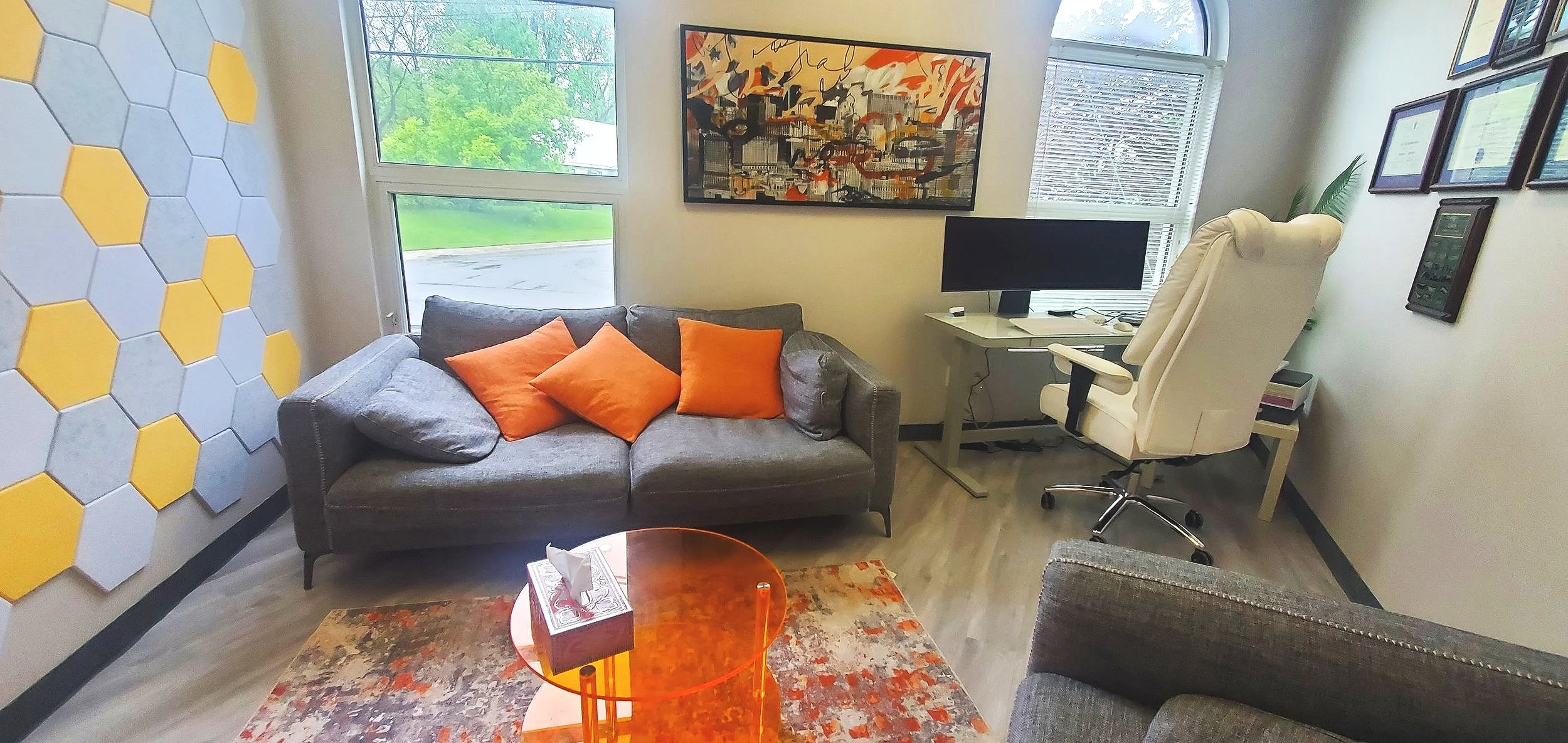 A cozy office with a gray sofa with orange pillows, a white desk with a computer monitor, and a beige high-back office chair. There are colorful abstract paintings on the wall, and a large window showing green trees outside. A patterned area rug and an orange glass coffee table are also visible.
