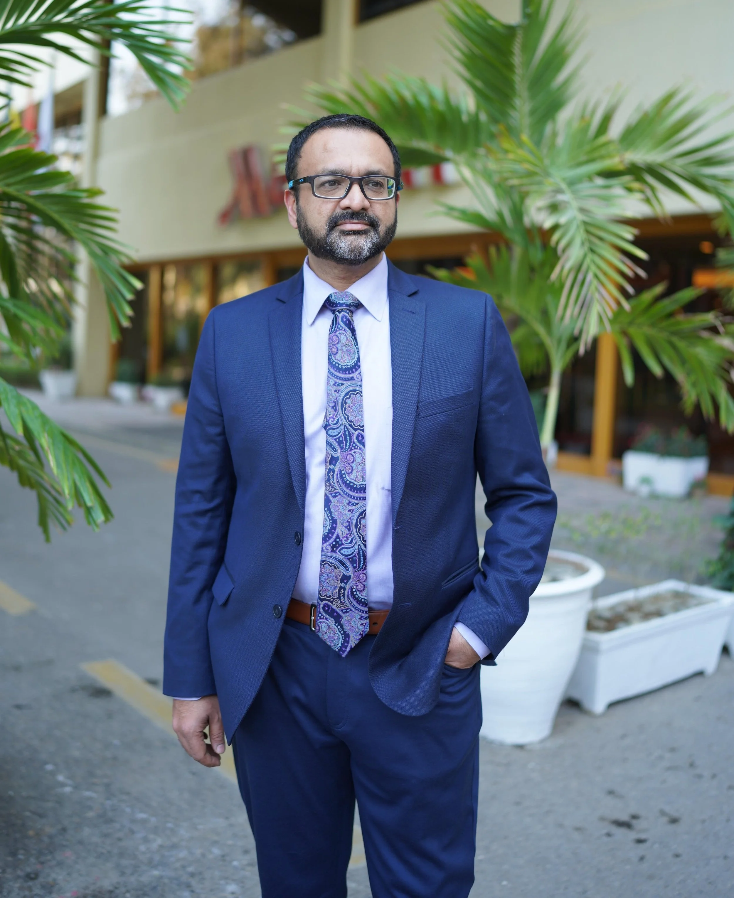 A man wearing a blue suit, white shirt, and paisley tie standing outdoors near green plants, with a building in the background.