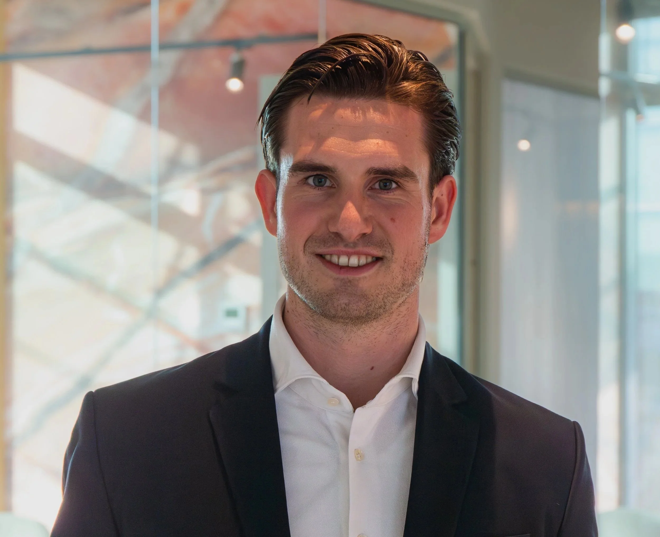 Portret van een glimlachende persoon met blond haar, gekleed in een zwarte jas, staande voor een glazen achtergrond in een bedrijfsomgeving.