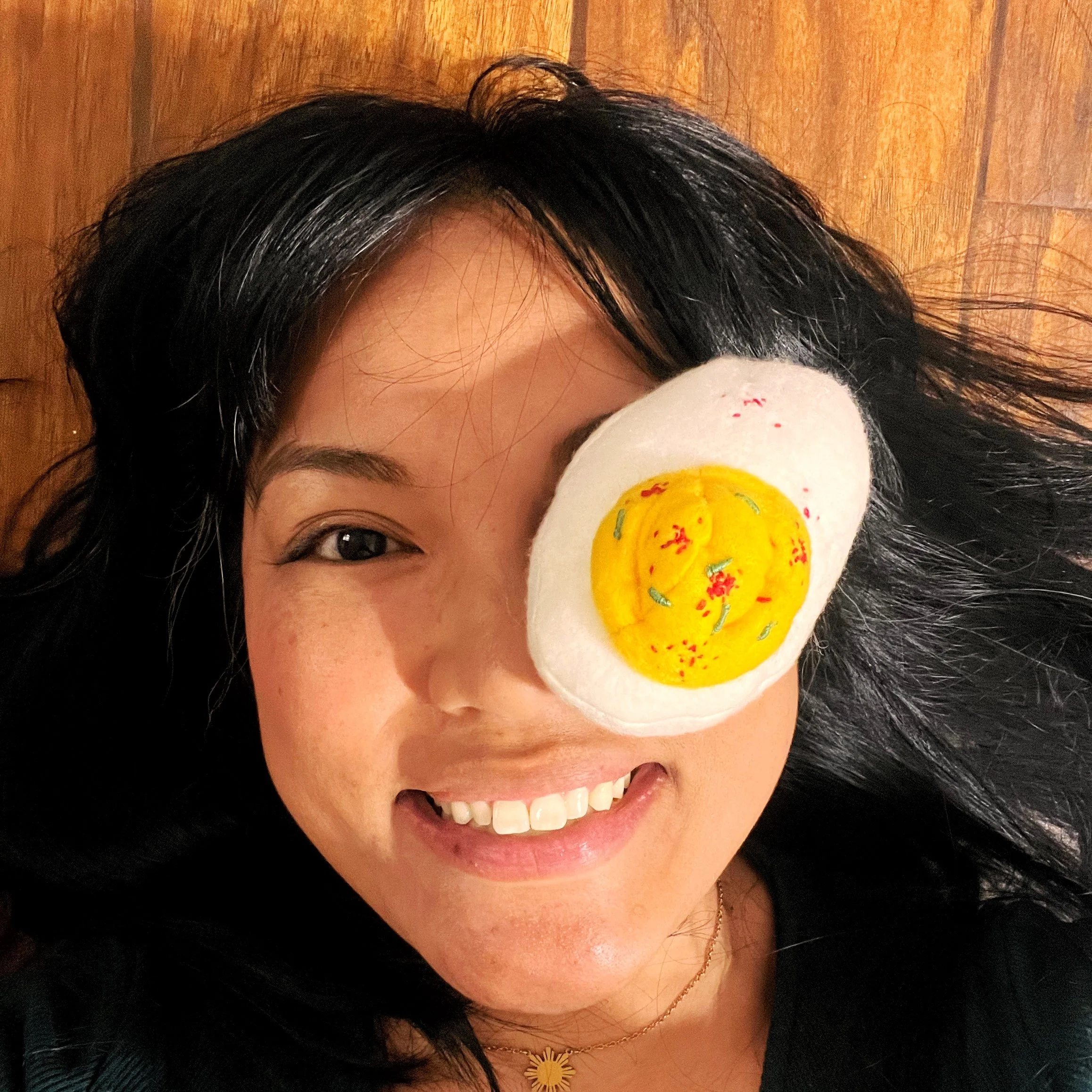Woman lying on a wooden floor with a plush object resembling a fried egg covering one eye, smiling with teeth visible.