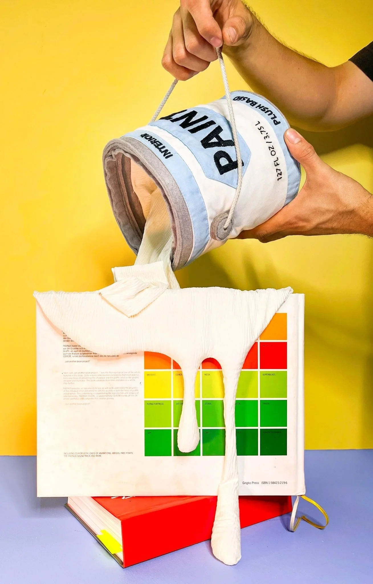 A plush paint can bucket with plush paint being poured out over a stack of books against a yellow and blue background.