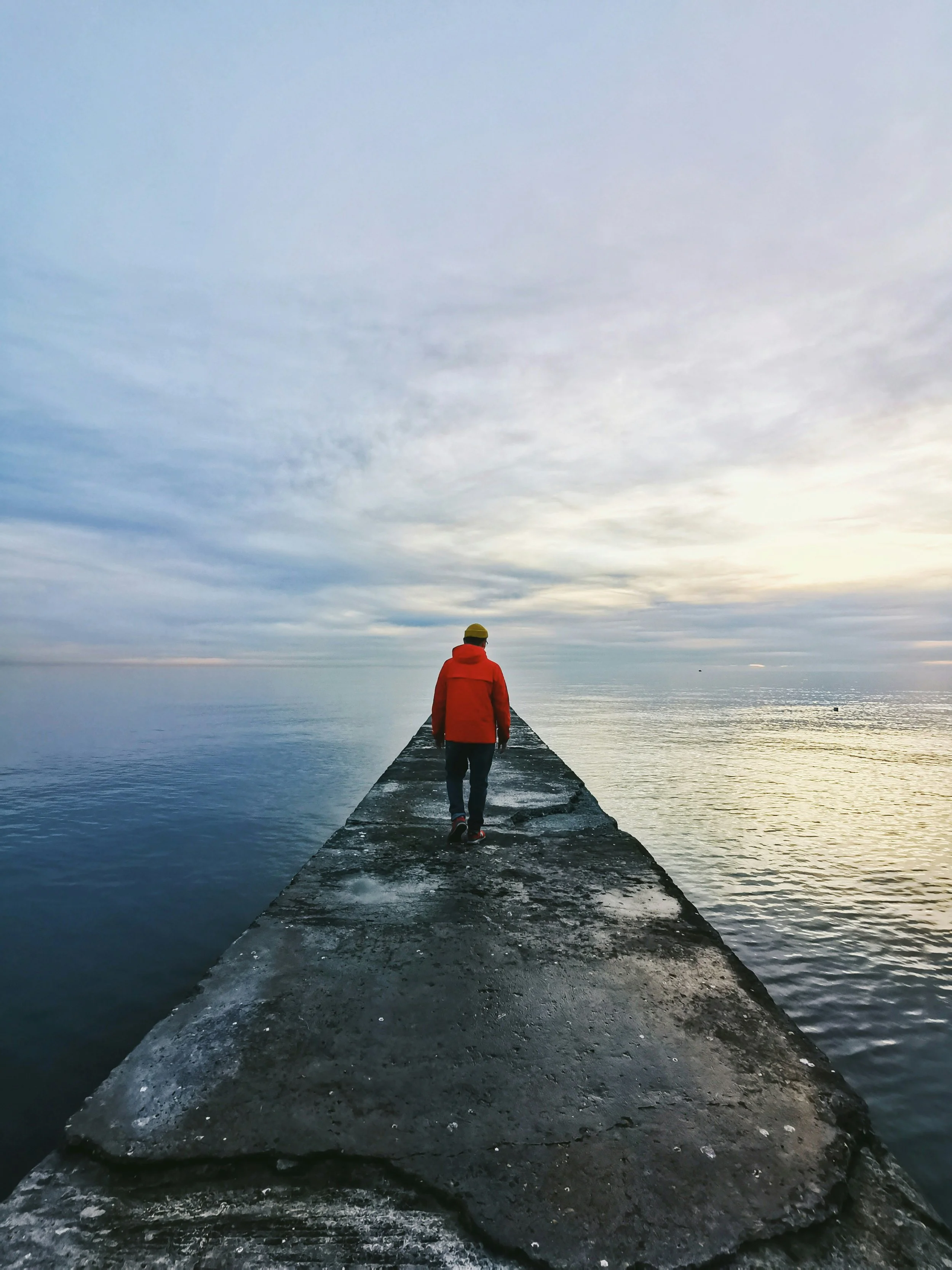 Person in red jacket walking on a pier over calm water with a cloudy sky.
