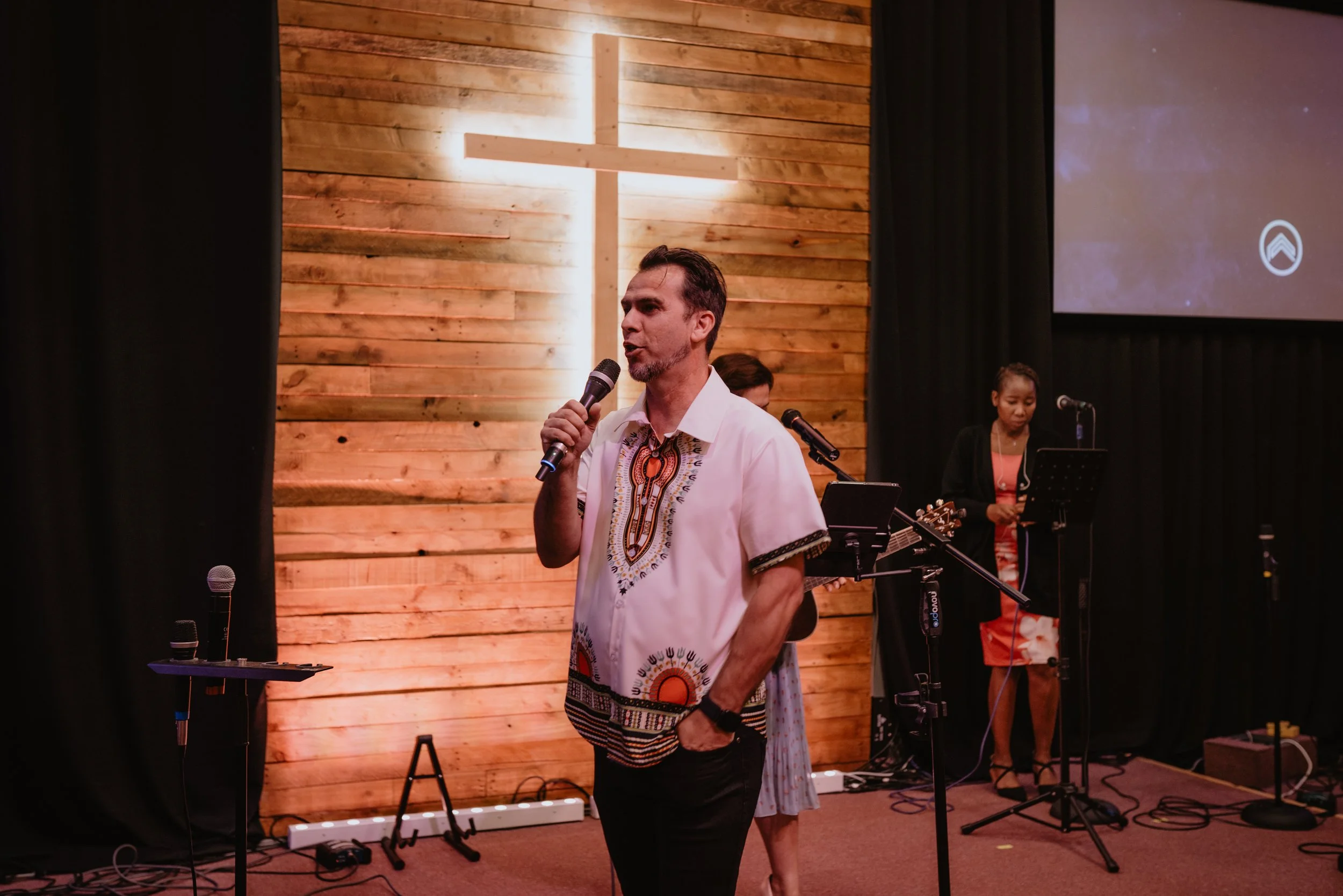 A man speaking into a microphone during a church service or event, with a wooden cross illuminated on a wooden wall behind him. Two women are in the background, one holding a microphone and standing at a music stand. The setting appears to be a church or worship space.