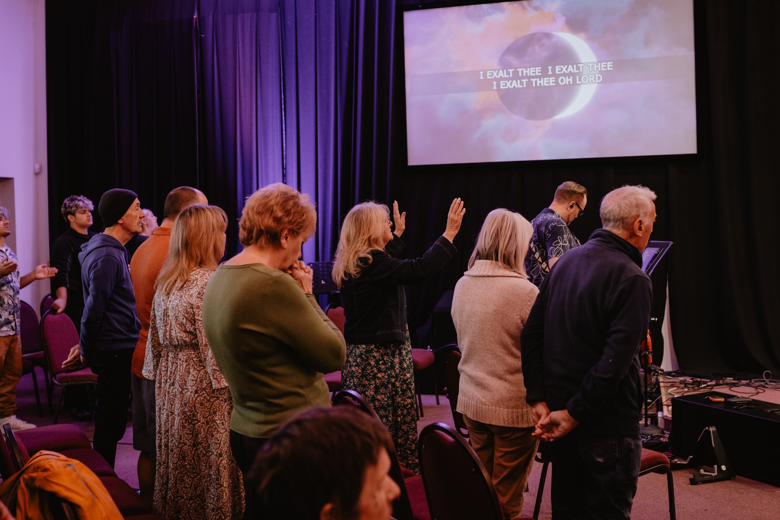 People standing with eyes closed or raised hands in a church worship service, with lyrics on a screen that read "I exalt thee, I exalt thee, I exalt thee oh Lord."
