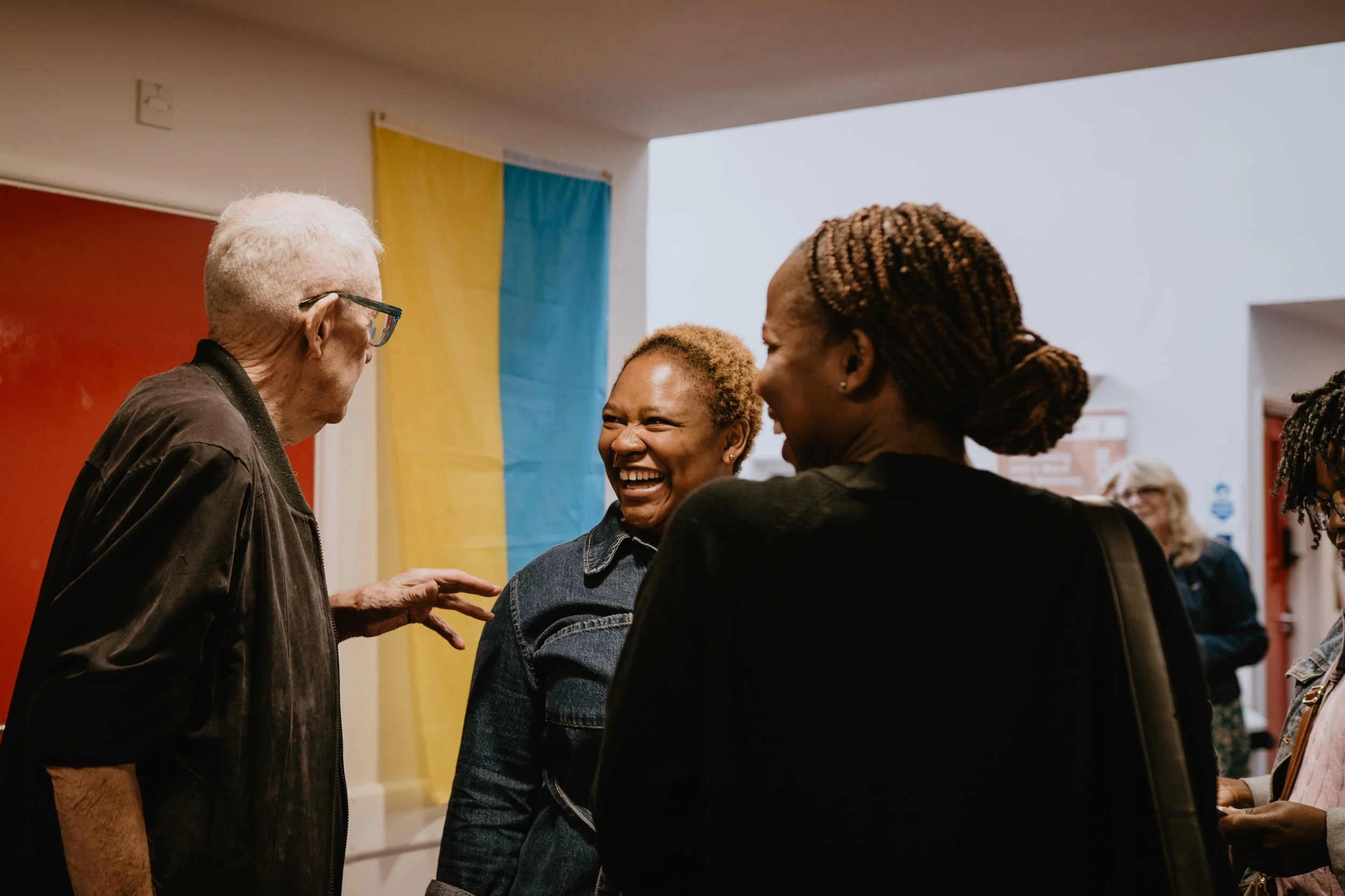 A group of four women, including one with gray hair and glasses, are laughing and smiling together indoors. The background features a yellow and blue flag, and additional people are visible in the distance.