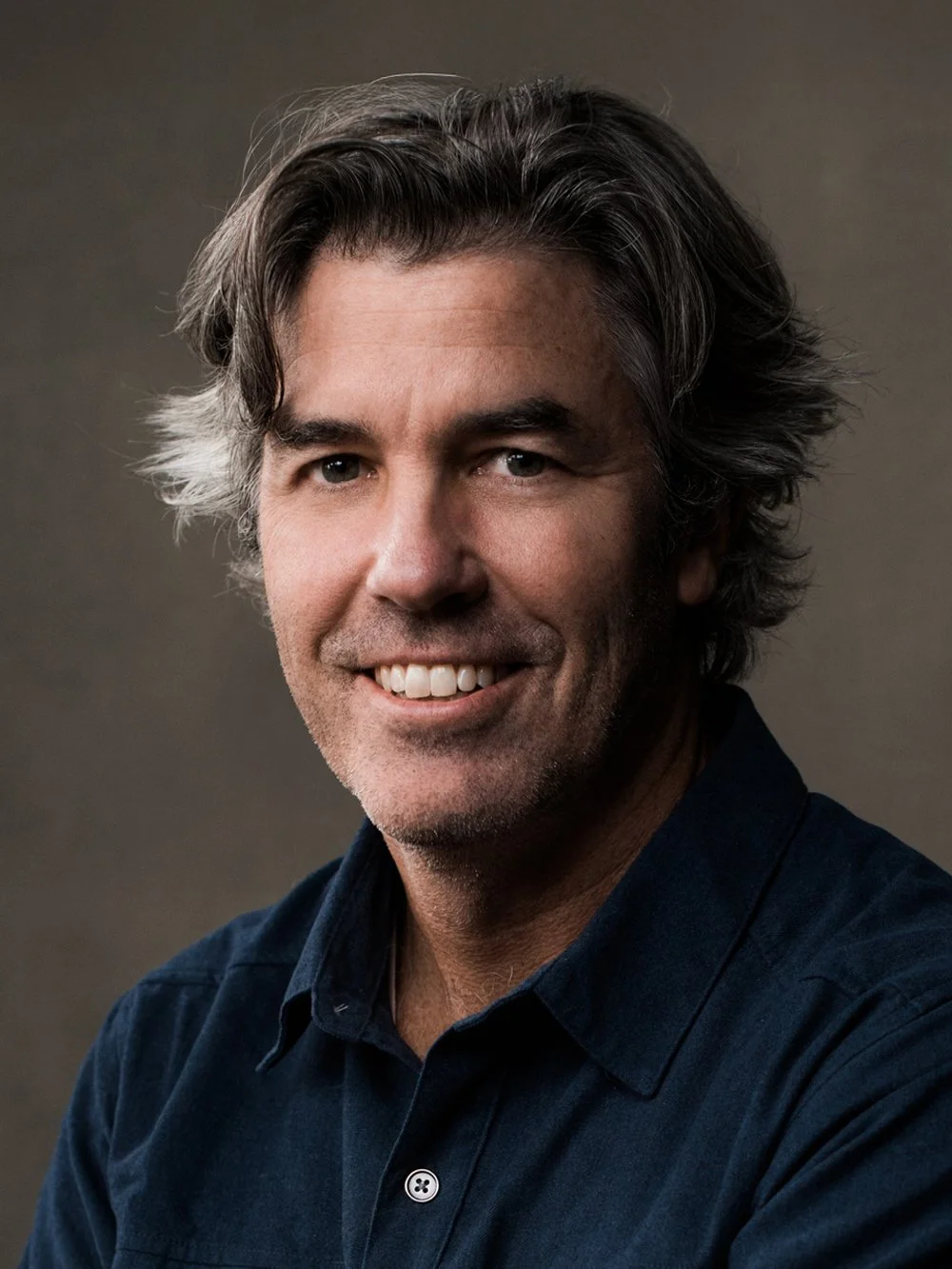 Portrait of a smiling man with wavy salt-and-pepper hair, wearing a dark button-up shirt, against a neutral background.