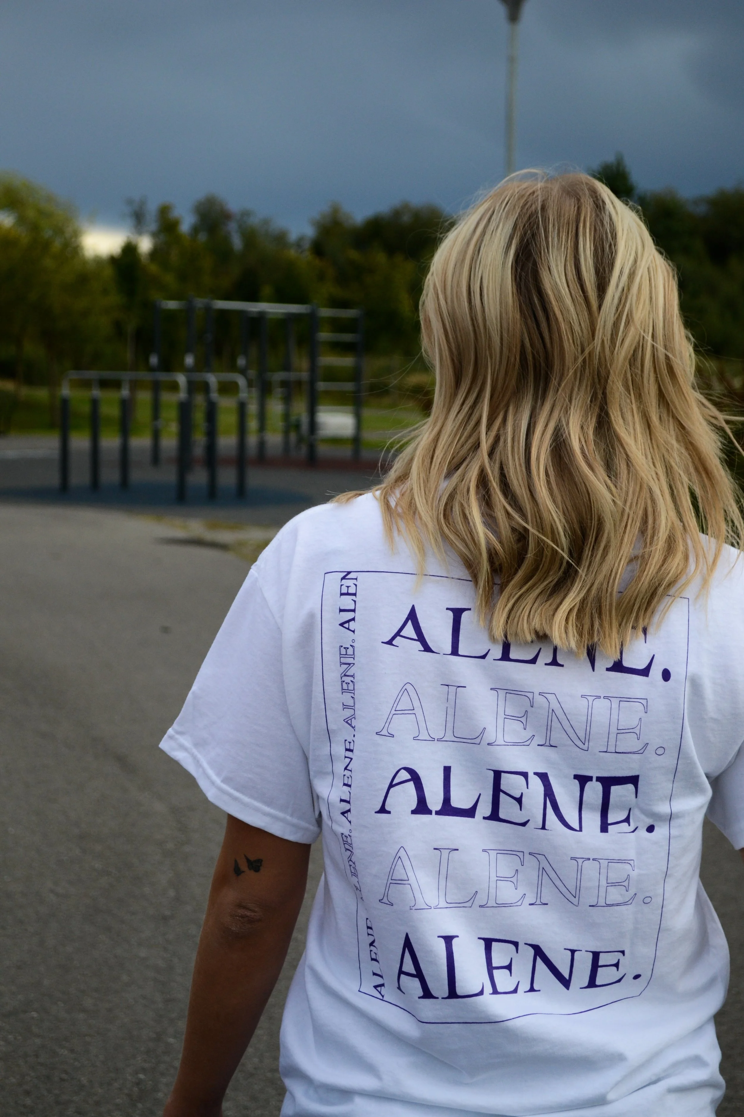 Person med langt blondt hår som vender seg bort, har på seg hvit T-skjorte med teksten 'ALENE' skrevet i ulike stiler. Person står utendørs i nærheten av en lekeplass med klatrestativer, under en mørk, overskyet himmel.