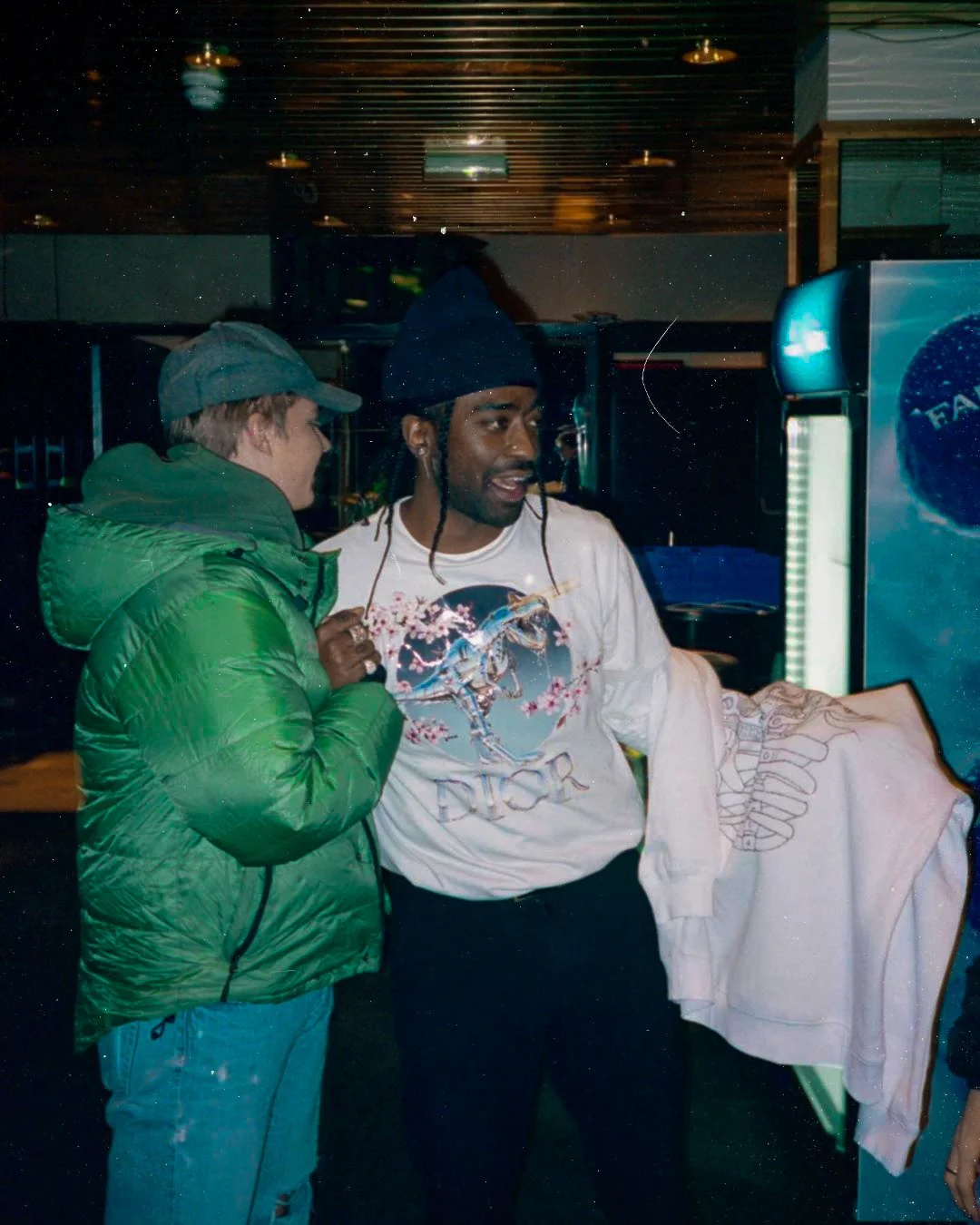 To personer står ved en frysedisk i en butikk, én person har på seg en grønn jakke og en grå caps, den andre har på seg en hvit Dior t-skjorte og en mørk lue. Begge ser ut til å snakke.