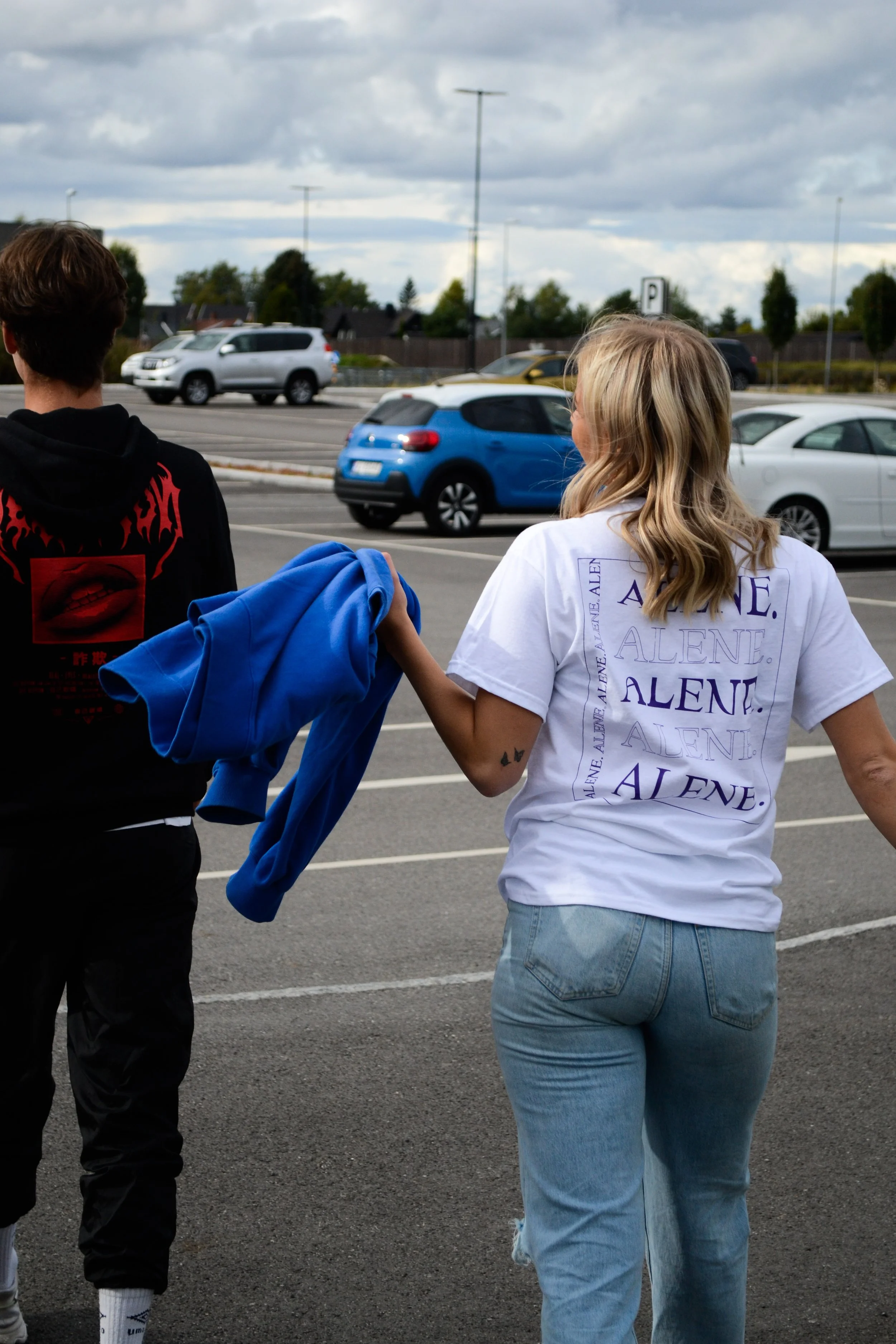 To personer går sammen i en parkeringsplass. Jente har på seg en hvit t-skjorte med teksten 'ALENE' skrevet i ulike stiler og farger. Gutt har på seg en svart hoodie med rødt design.