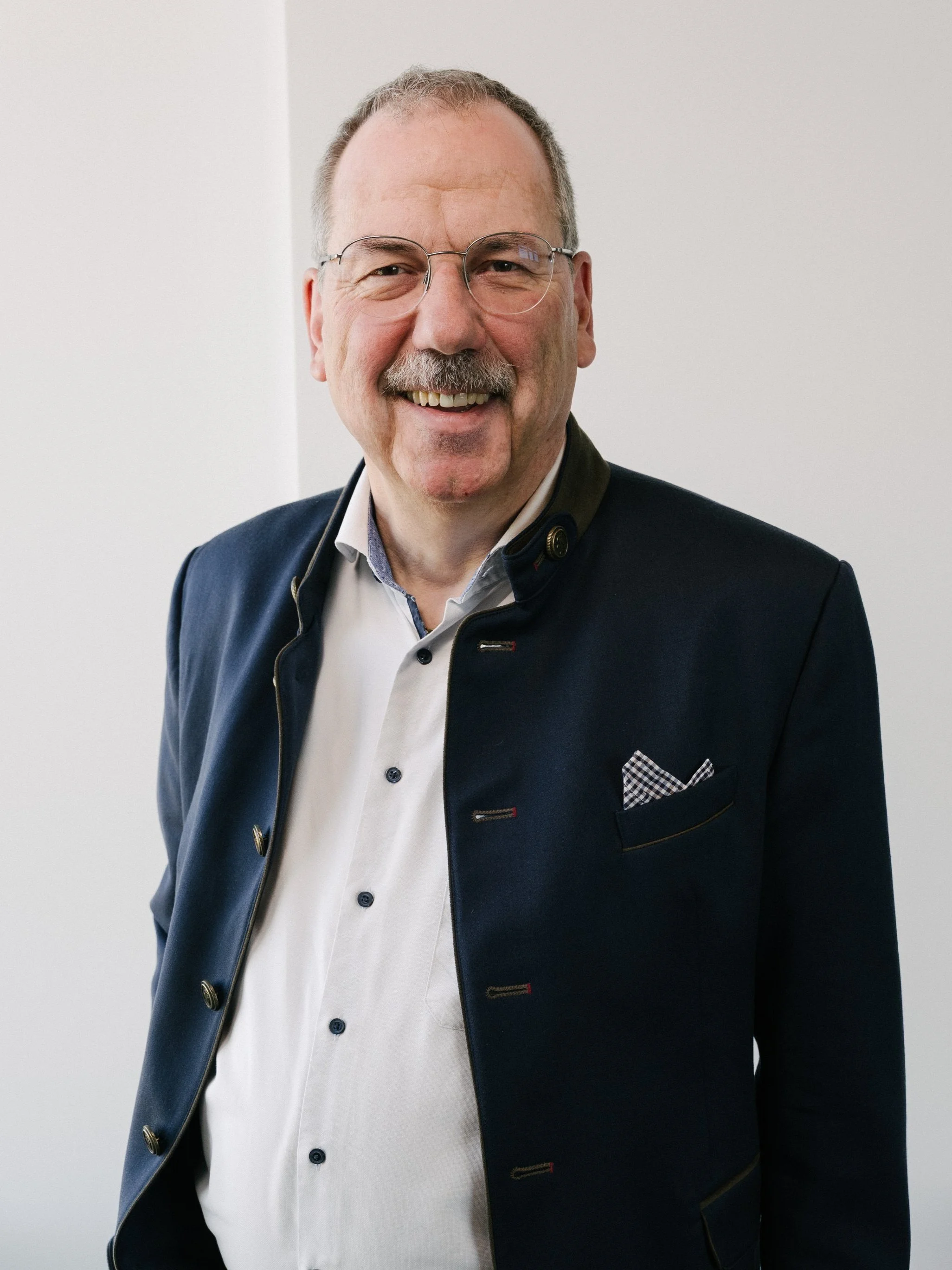 Portrait eines älteren Mannes mit Brille und Schnurrbart, der in die Kamera lächelt. Er trägt einen dunklen Blazer und ein weißes Hemd.