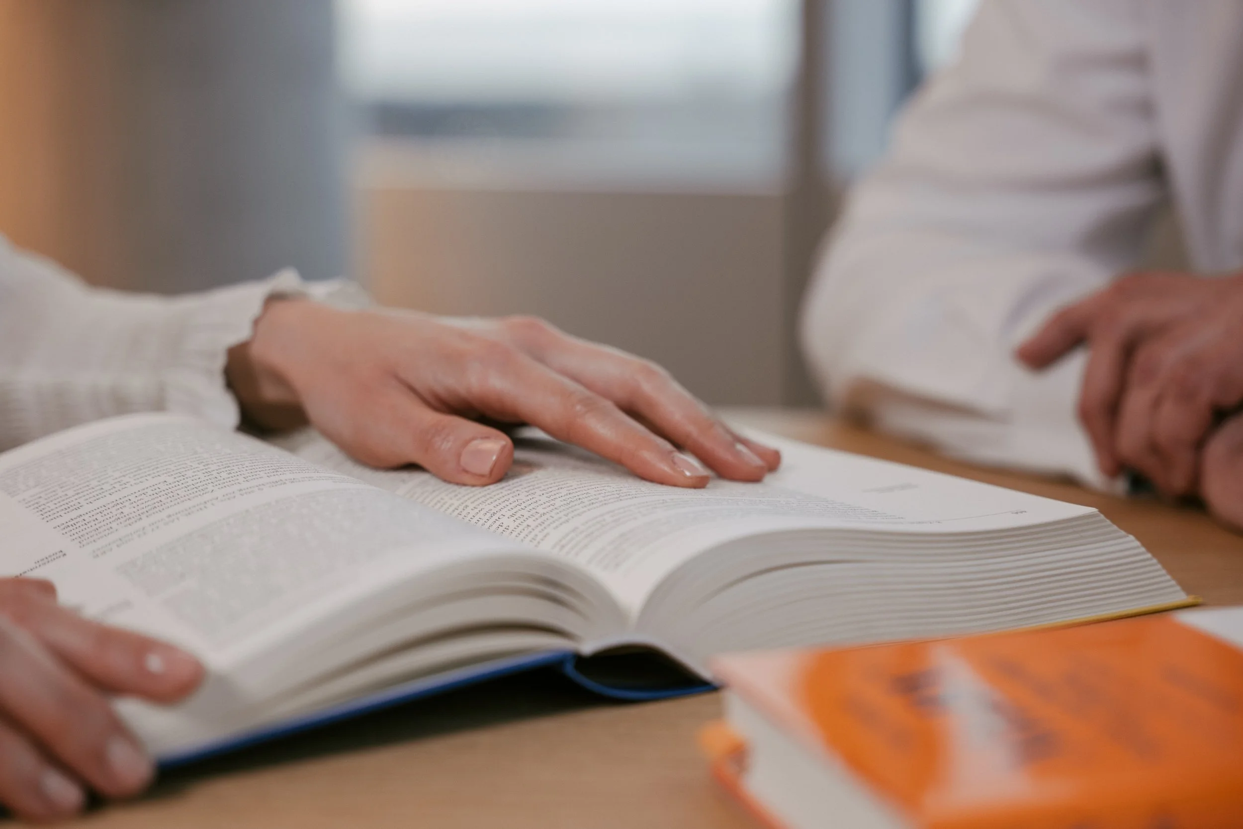 Nahaufnahme von zwei Personen, die an einem Tisch sitzen, mit einem offenen Buch. Eine Hand berührt die Buchseite.